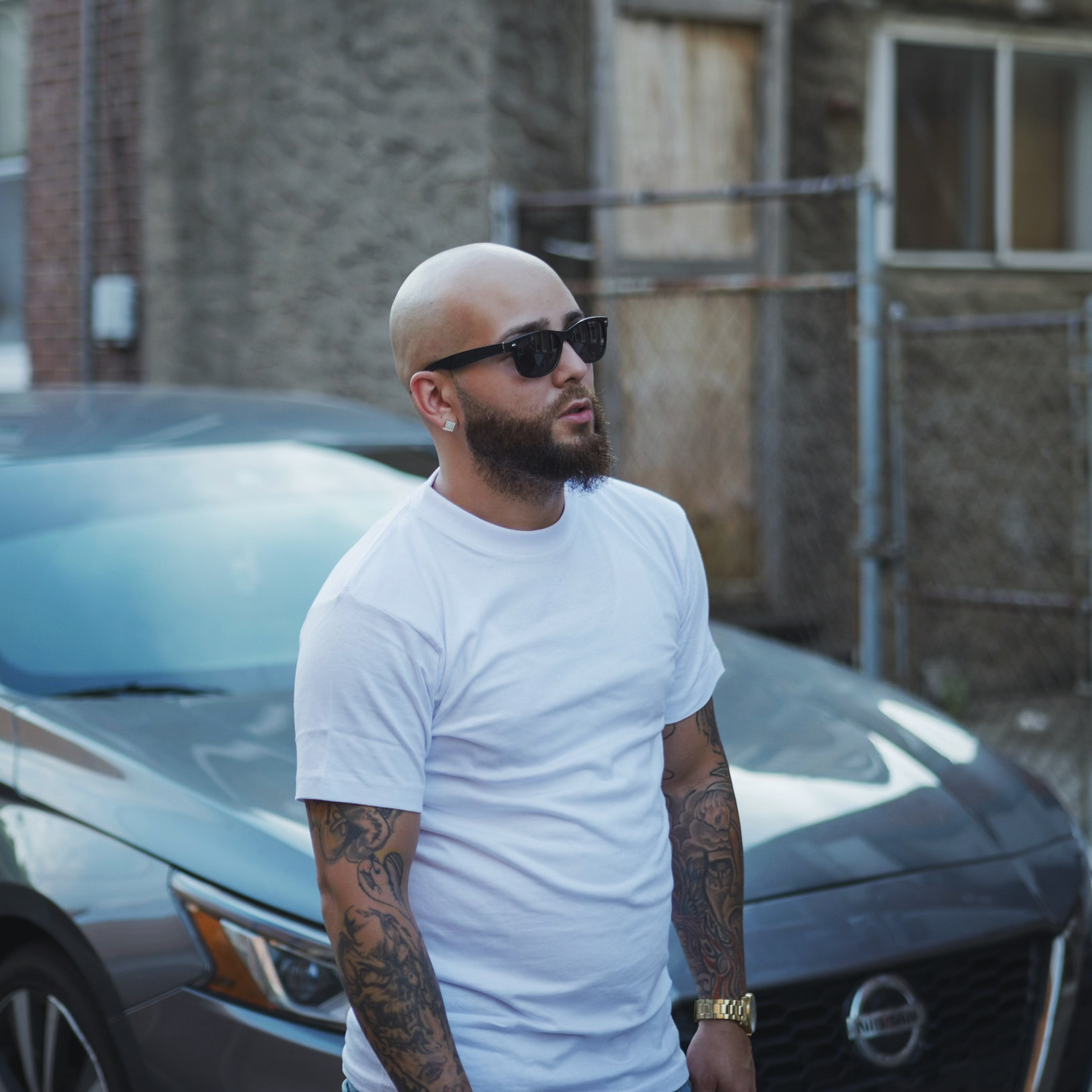 Bald man with tattoos wearing sunglasses and a white t-shirt standing outdoors near cars and a building.