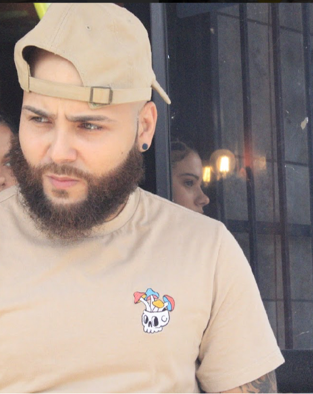 Man with beard, wearing a beige cap backward and a beige T-shirt with a skull and rainbow patch, looking to the side.
