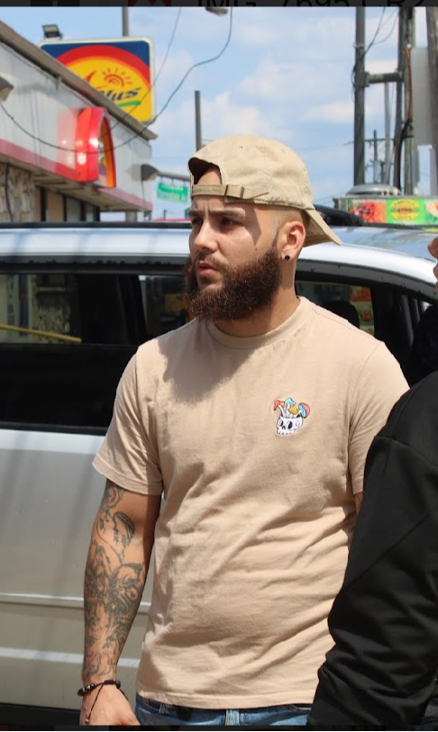 Man with beard, tattoos, beige cap, and beige T-shirt standing outdoors near cars, with signs and power lines in background.