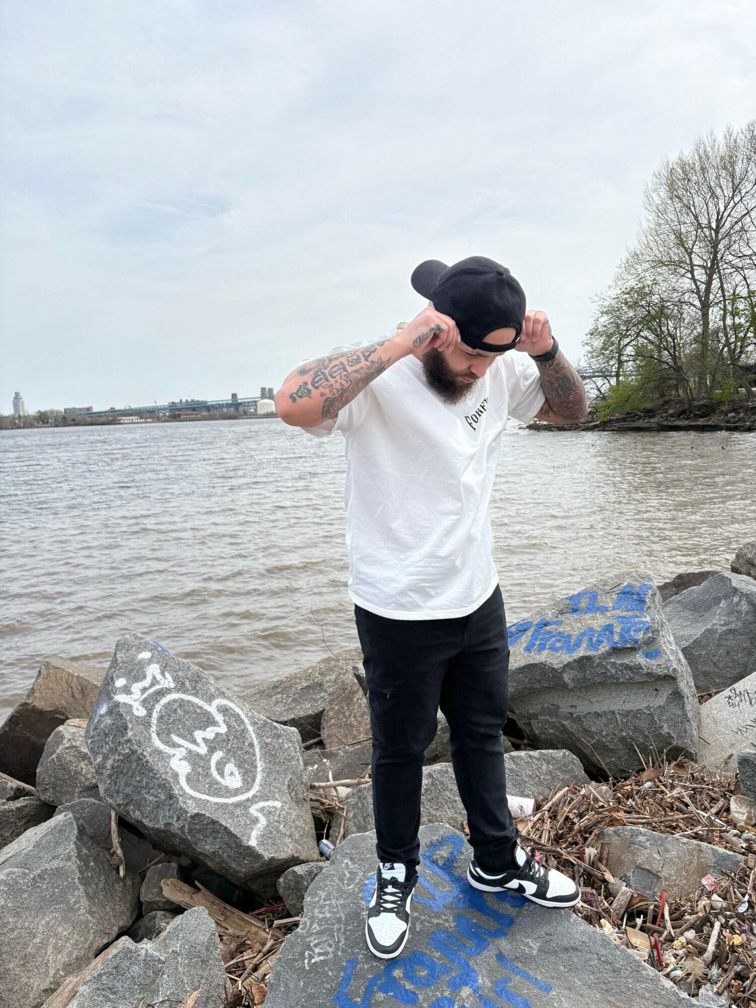 Person standing on rocks near water, talking on phone, wearing sunglasses, white shirt, black pants, and sneakers.