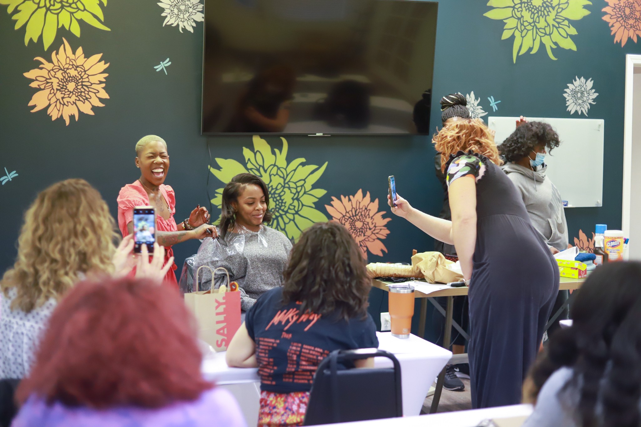 Group of women and children in a room with floral wall decor, some taking photos, others smiling and talking.