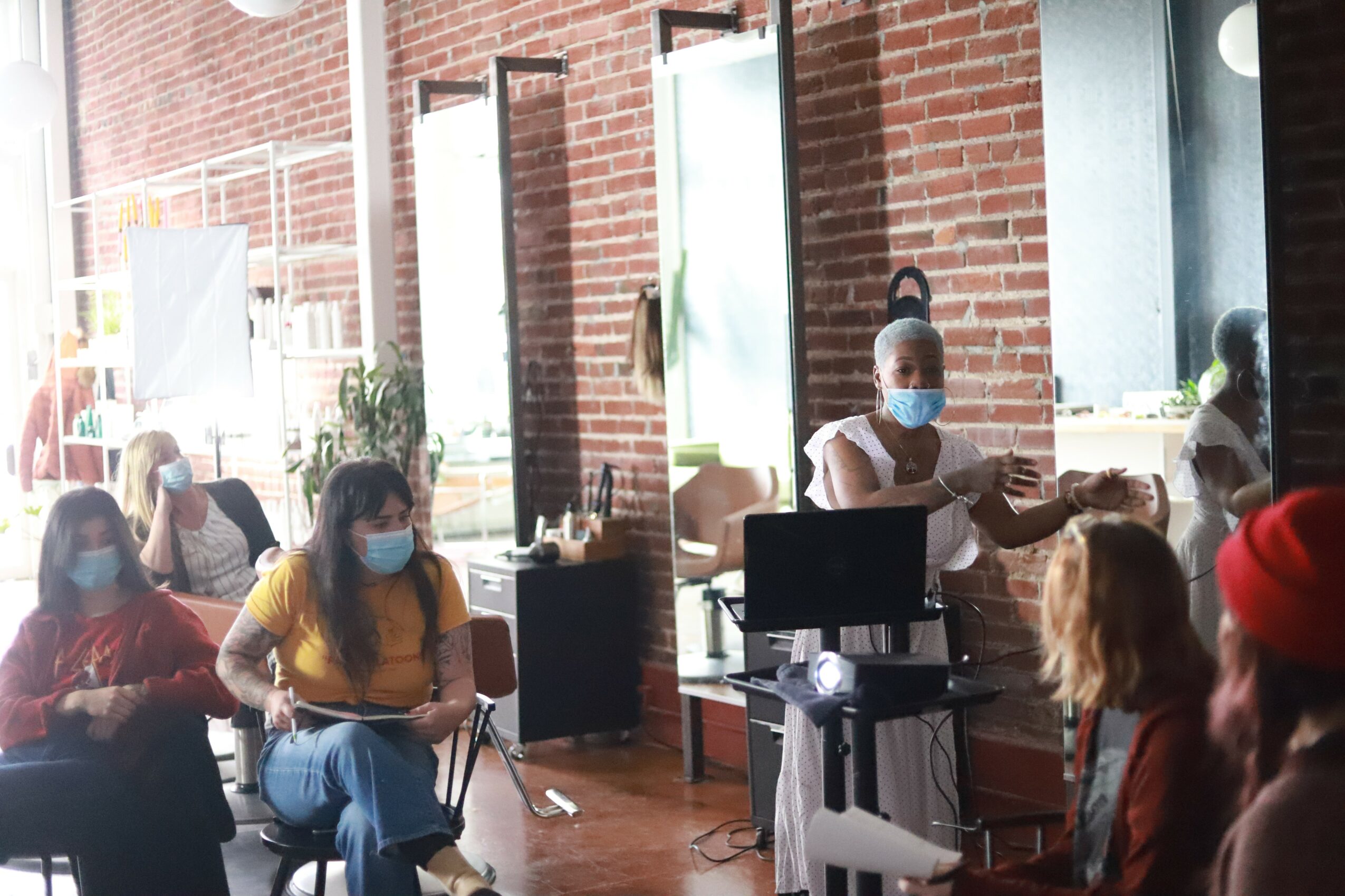 Group of people wearing masks sitting indoors, some engaging with a person at a desk, brick wall background.