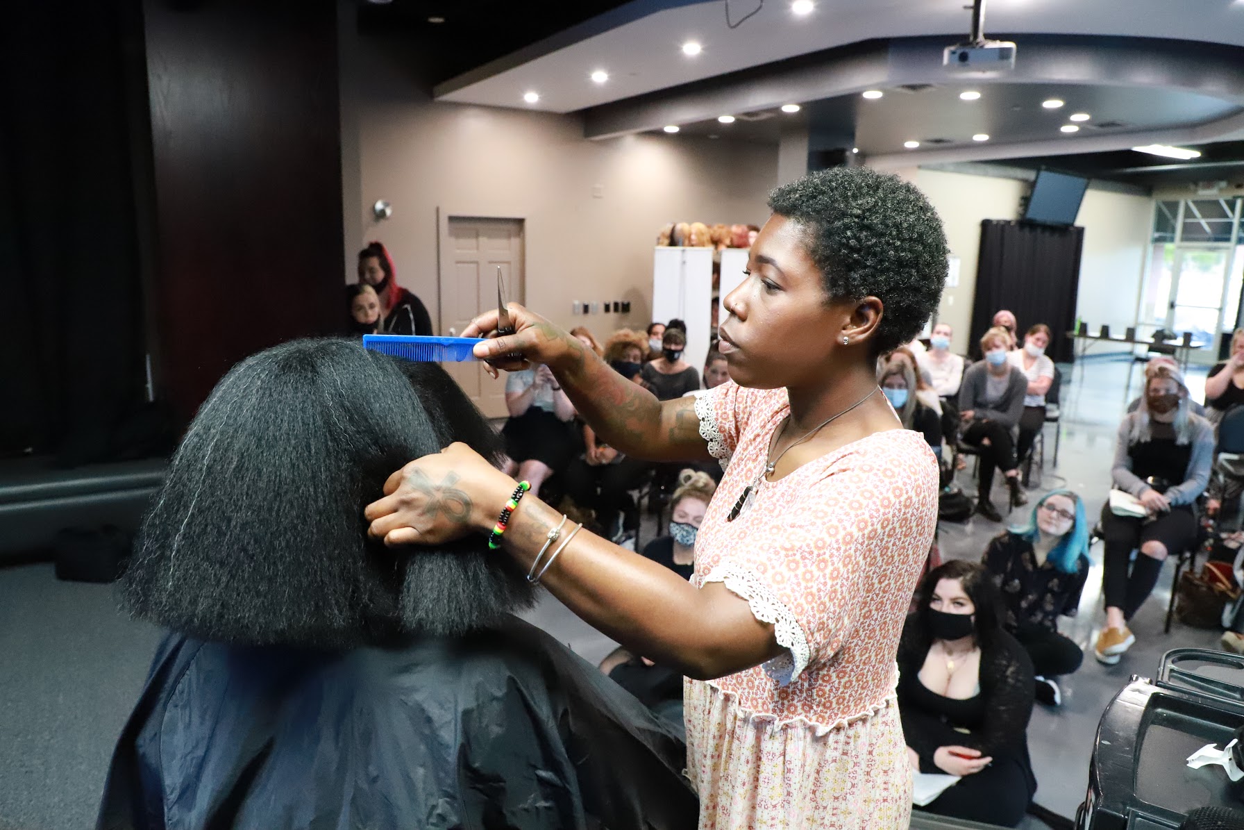 A woman styles a person's hair with a blue comb in a busy salon or event space.