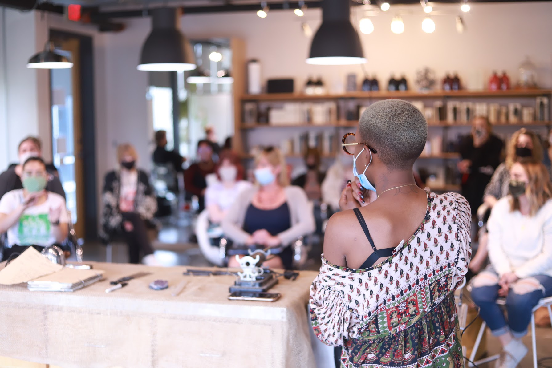 A woman with short hair and glasses wearing a face mask and patterned top, facing a group of people in a cafe or restaurant.