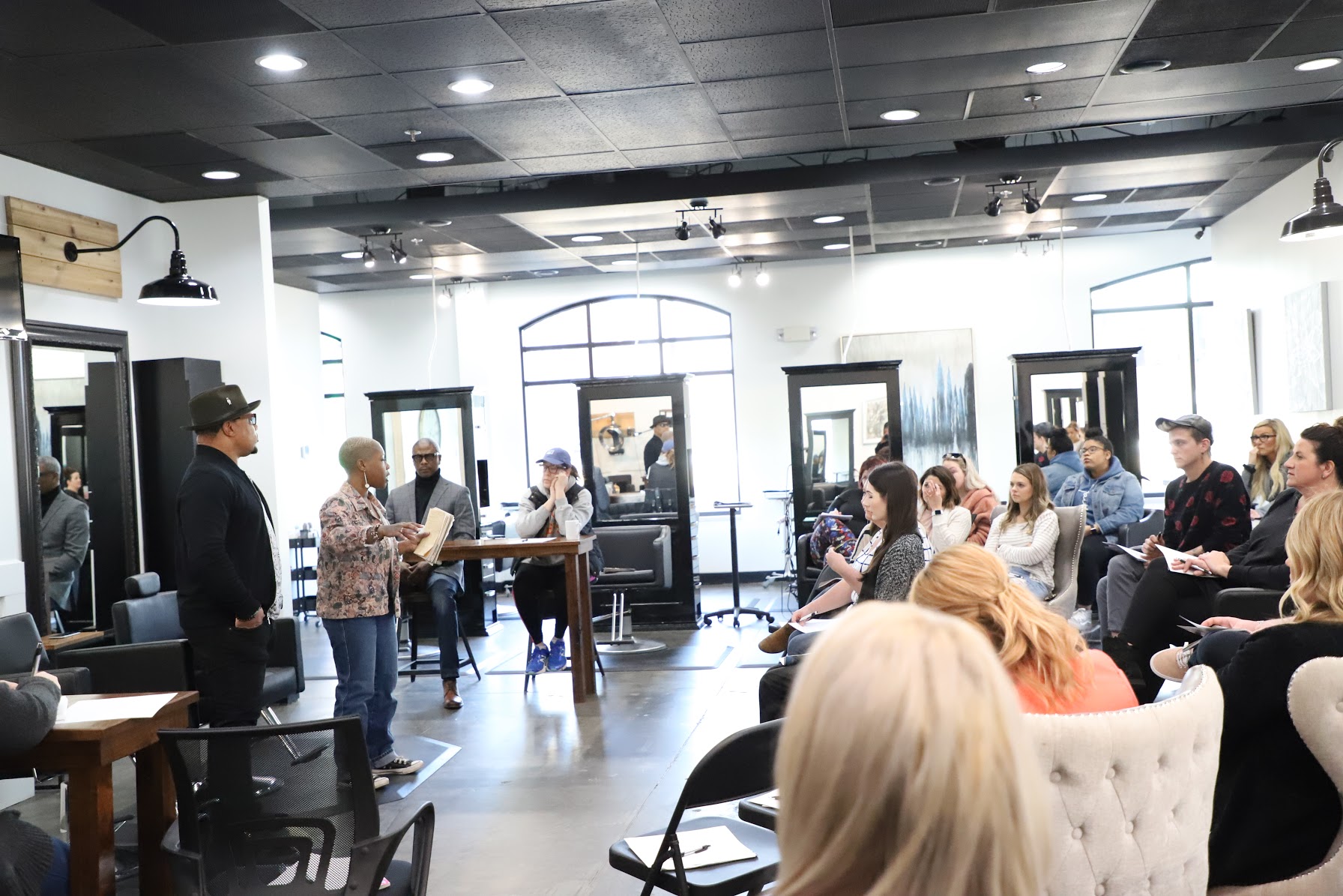 People gathered in a bright, modern room with large windows, listening to a speaker at the front, some seated and some standing.
