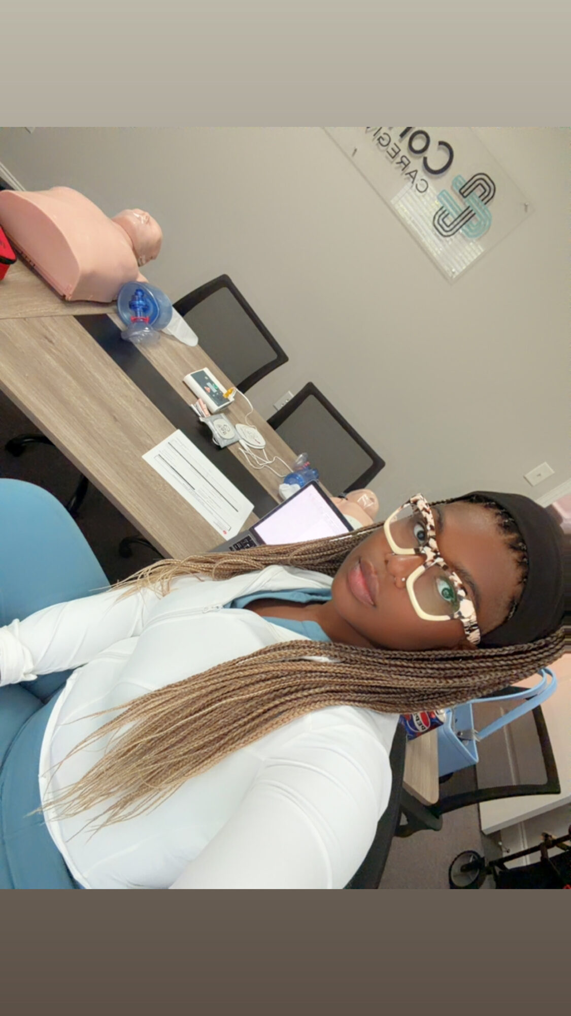 Woman with long hair and glasses sitting at a desk in an office, with computers and a poster on the wall.