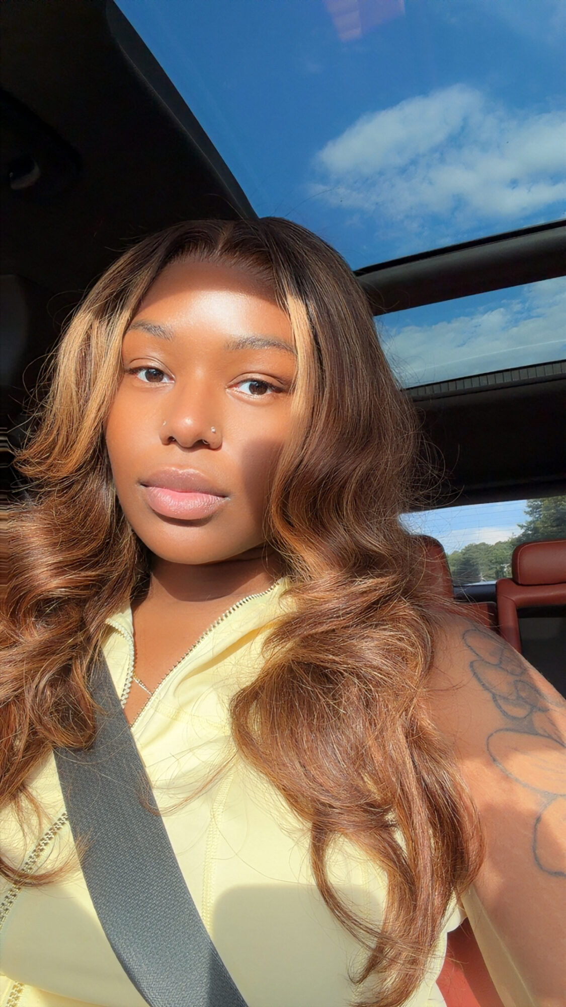 Young woman with long wavy hair sitting in a car, sunlight on her face, blue sky visible through sunroof.
