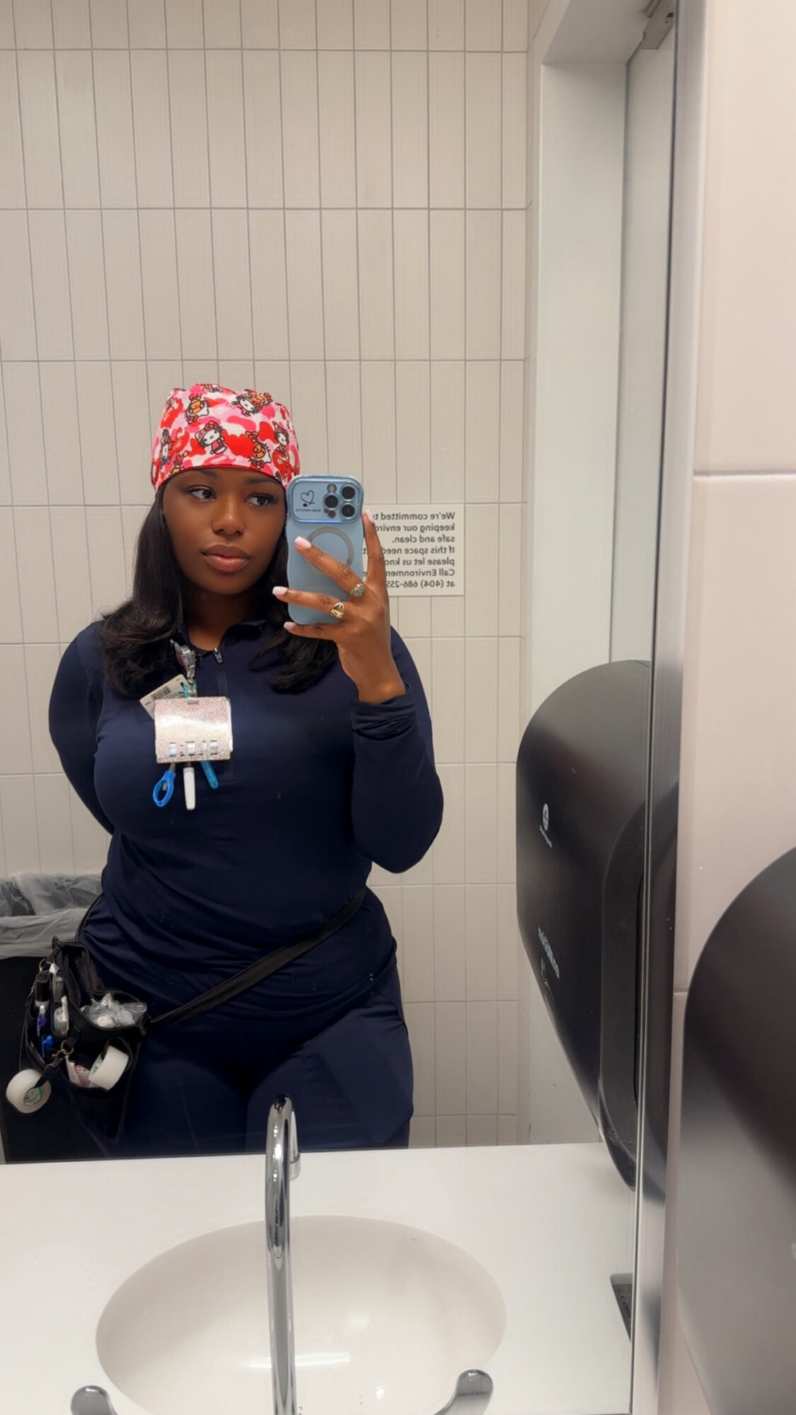 Person taking a mirror selfie in a bathroom, wearing a colorful headscarf and dark clothing, with a sink and mirror visible.