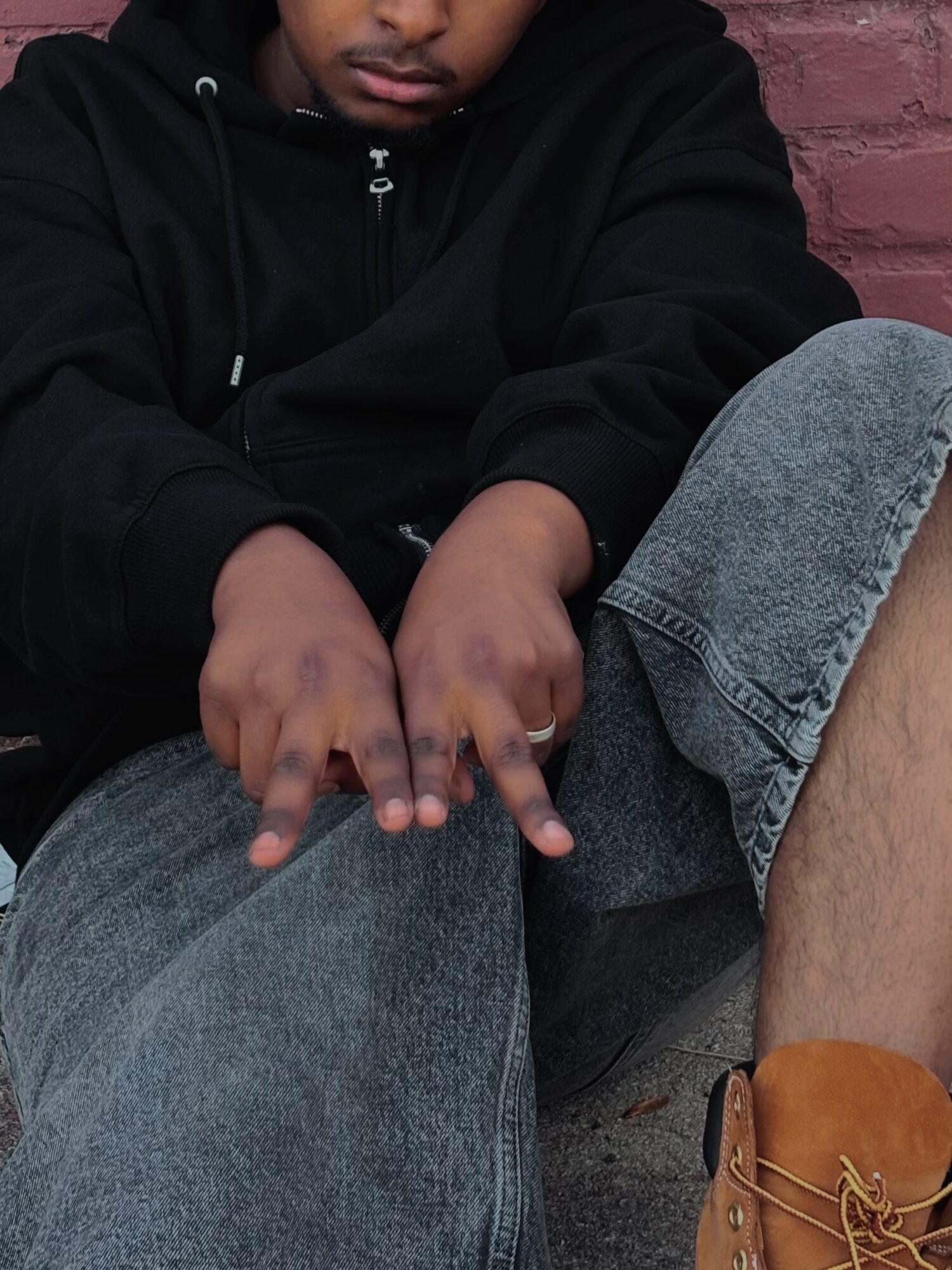 Person sitting outdoors, wearing a black hoodie and gray jeans, with hands resting on knee, against a brick wall background.