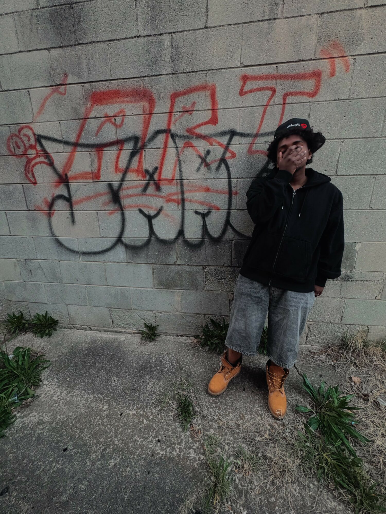 Person standing against a graffiti-covered wall, wearing a black jacket, gray shorts, and tan boots, with plants nearby.