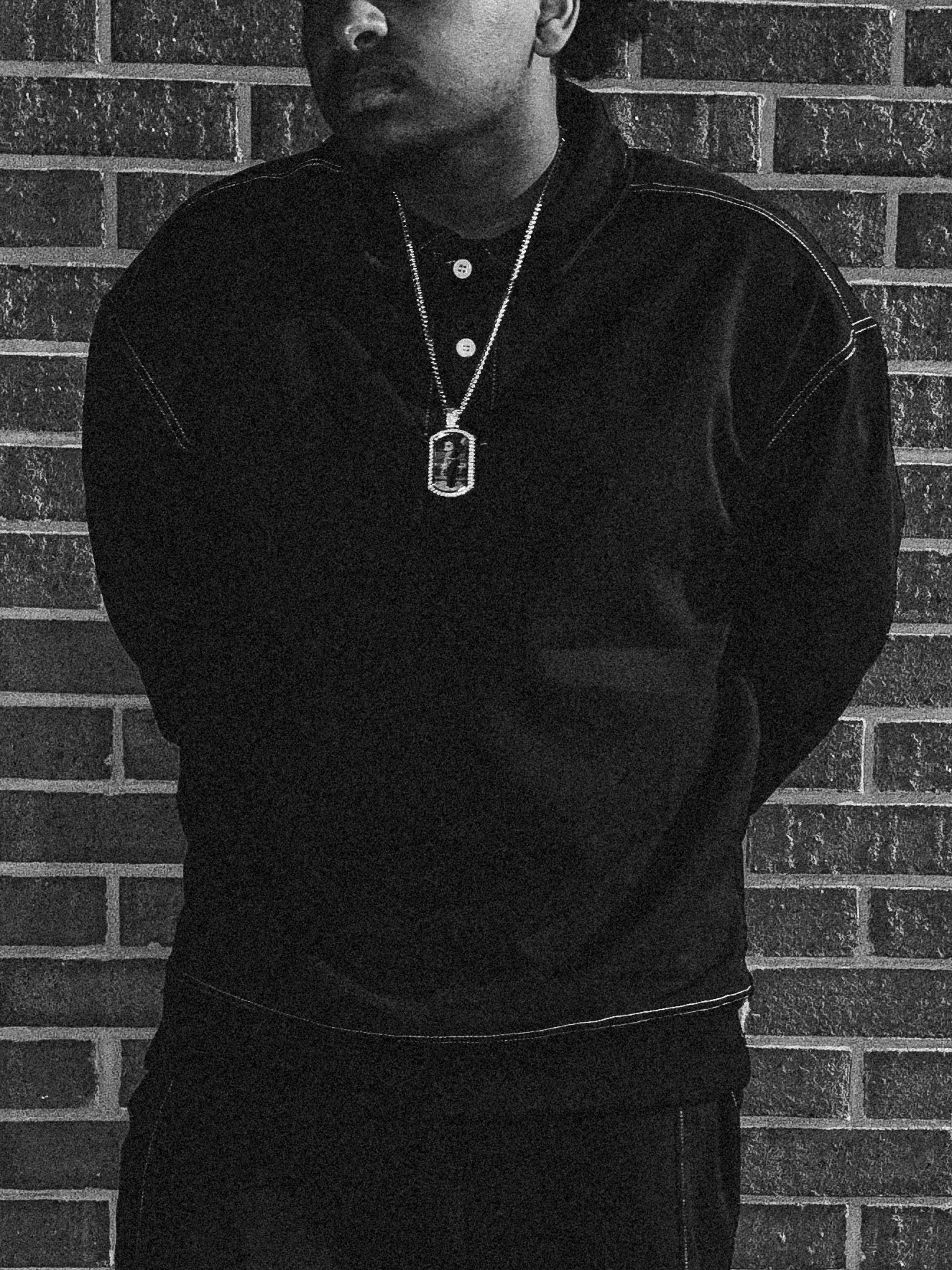 Person standing against a brick wall, wearing a dark top and a necklace with a pendant.