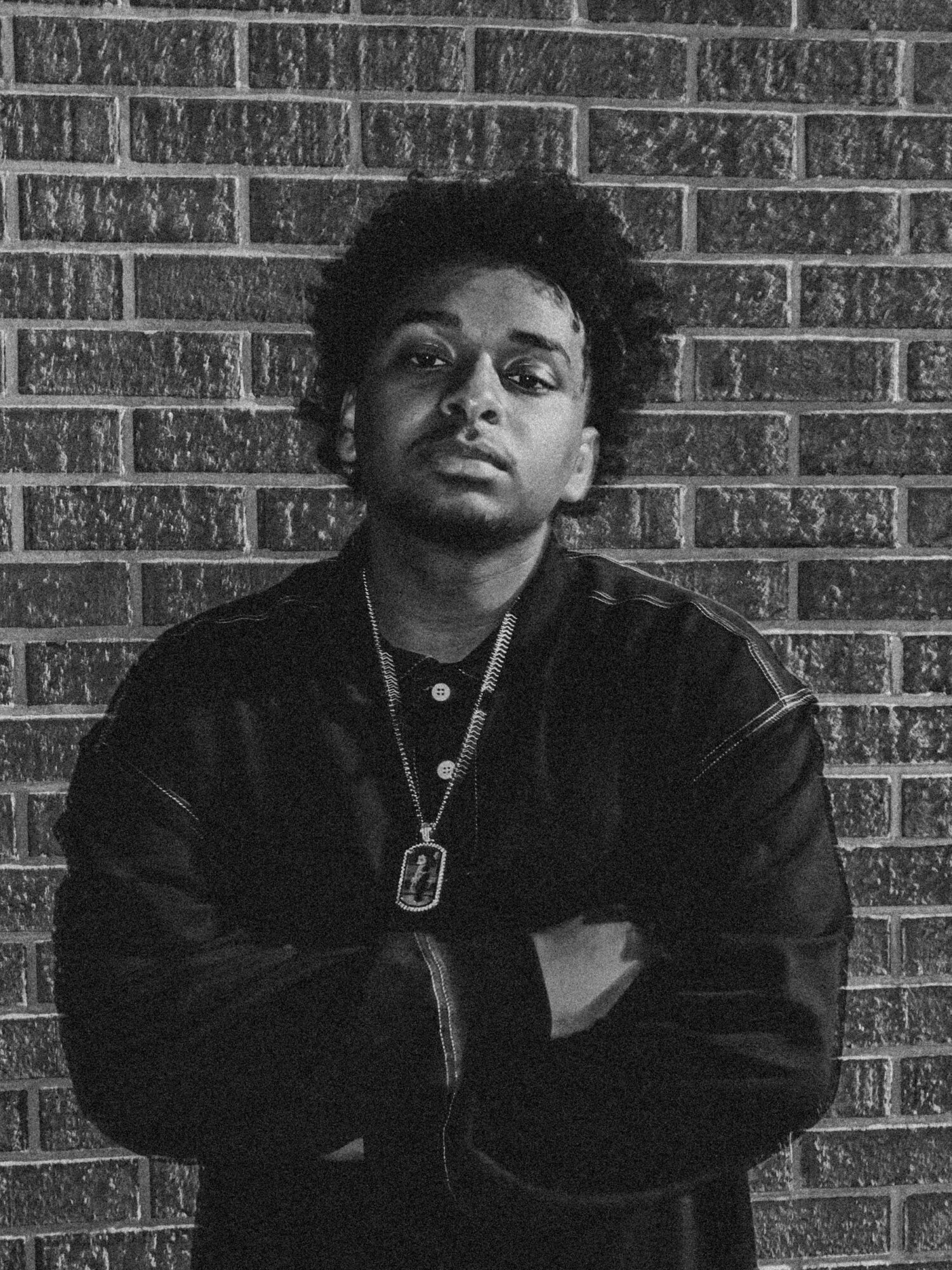 Person with crossed arms standing against a brick wall, wearing a jacket and necklace, looking at the camera.
