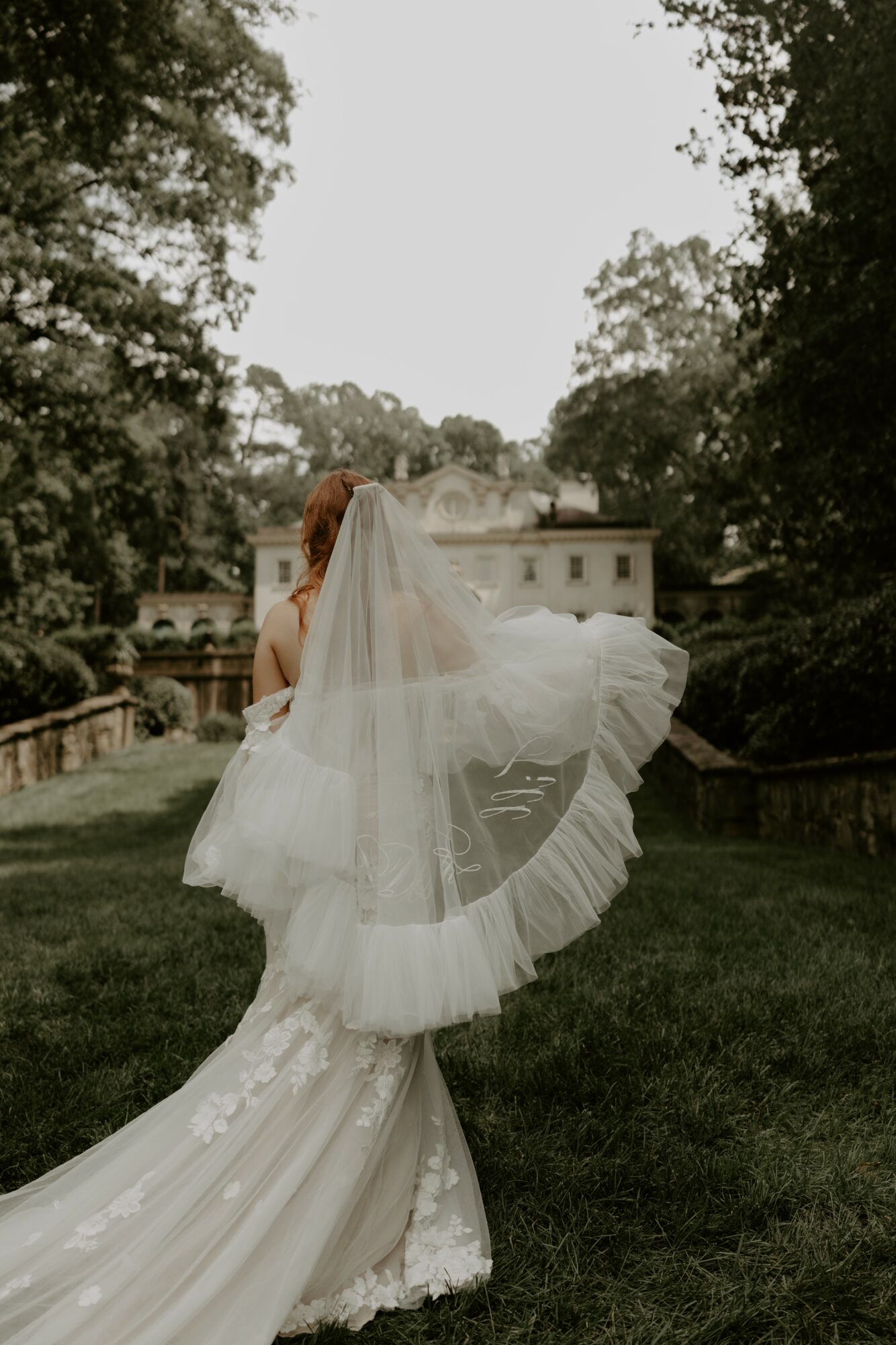 Bride in wedding dress and veil walking away on grass with trees and building in background.
