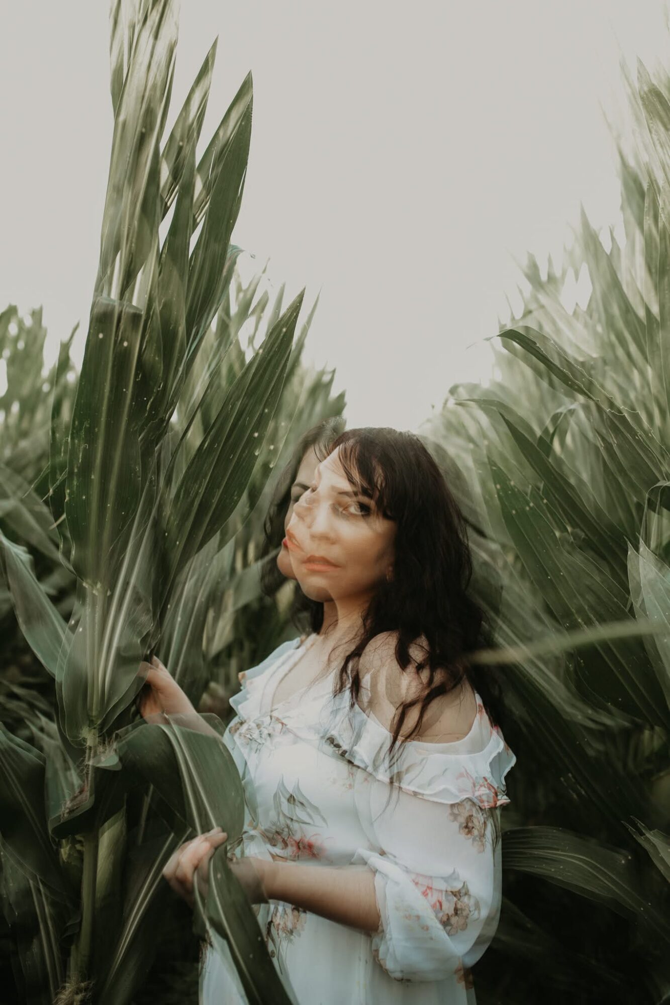 Woman with dark hair and glasses standing among tall green plants outdoors.