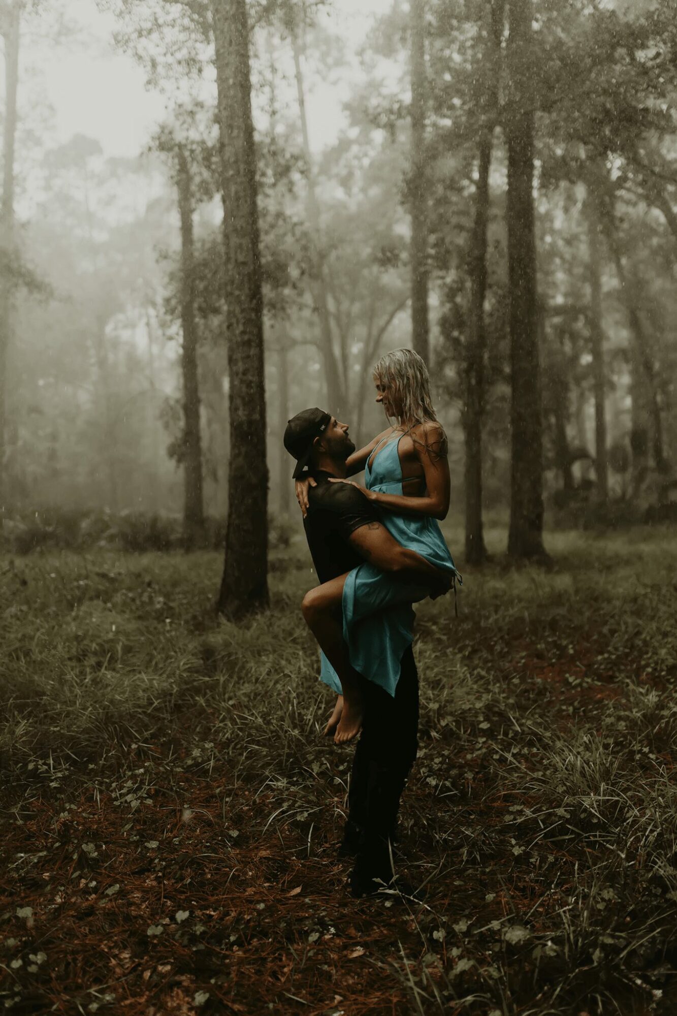 Two people in a forest, one lifting the other, surrounded by trees and fog, with green grass and brown ground.