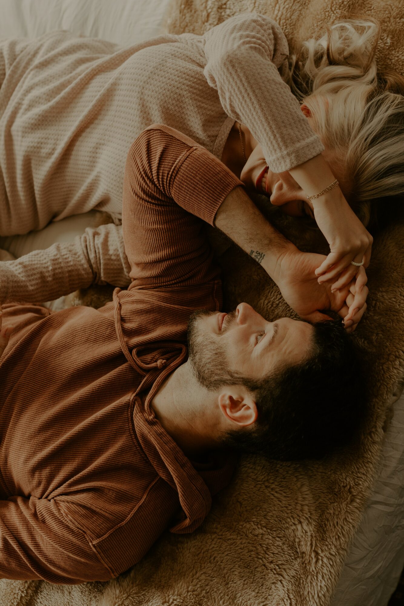 Two people lie on a bed, facing each other, holding hands, with relaxed expressions, in warm clothing.