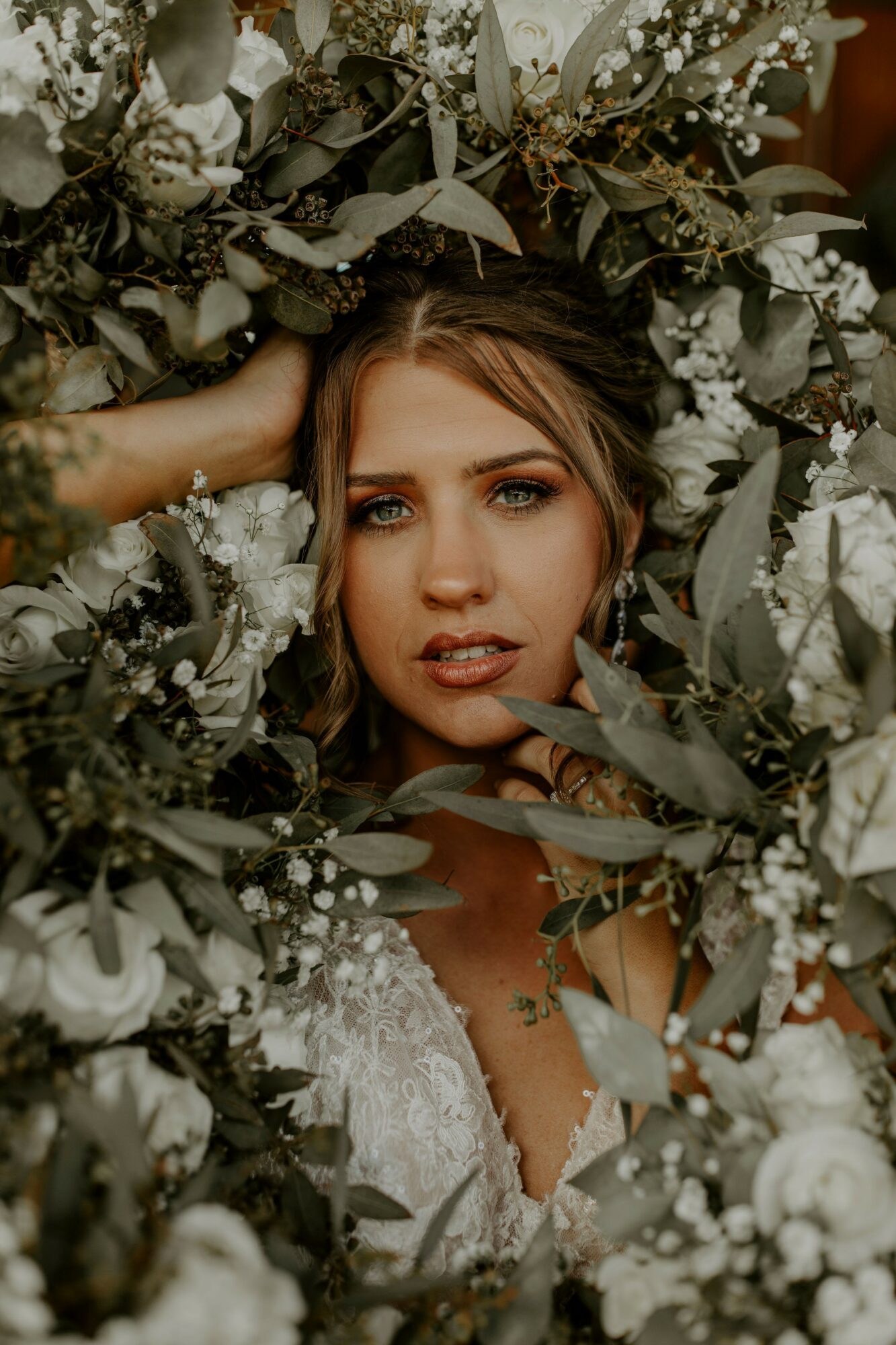 Woman with makeup and hair surrounded by white flowers and greenery, looking at the camera.