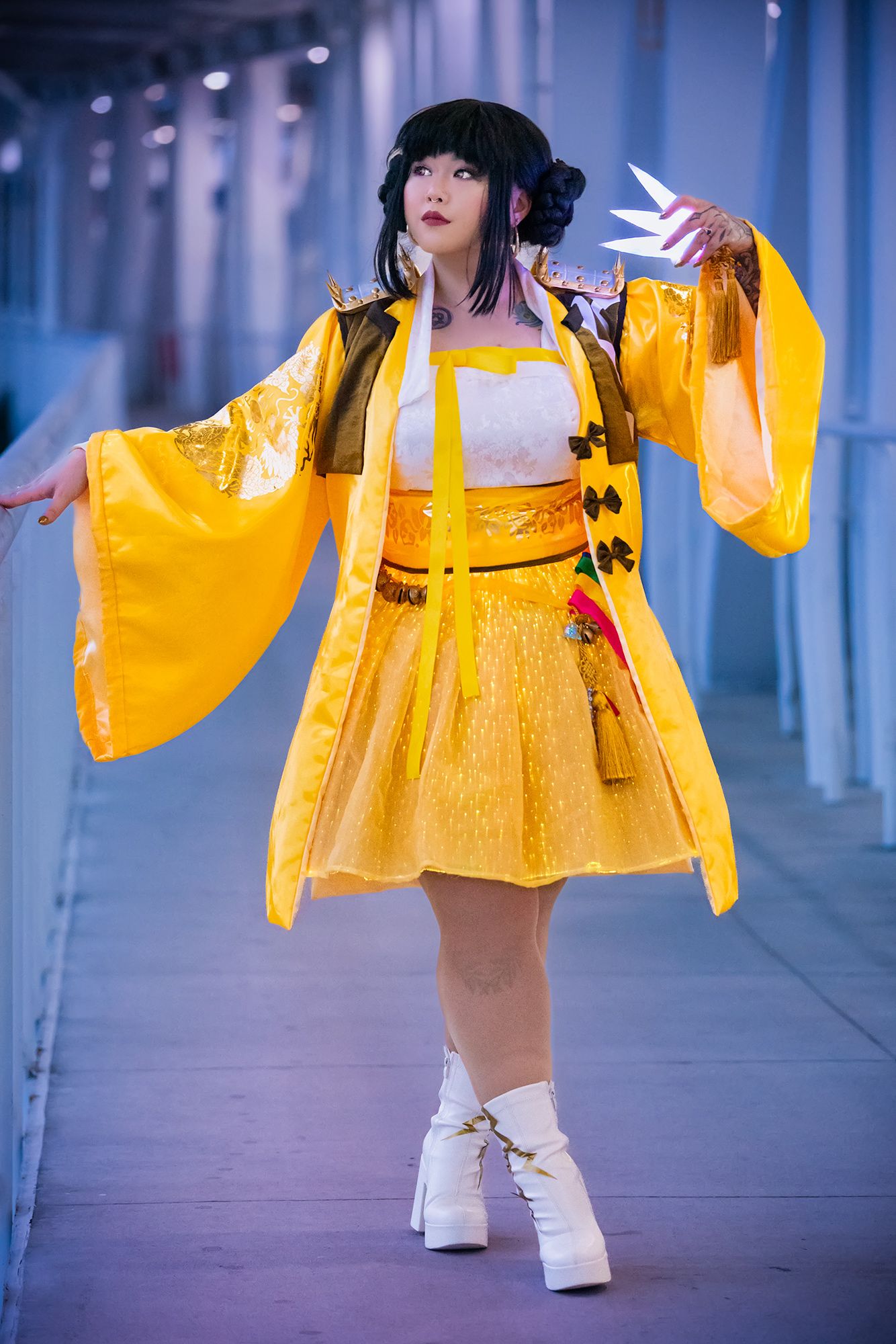Person in yellow costume with large sleeves, standing in a blue-lit corridor, holding a paper fan, with black hair styled in pigtails.