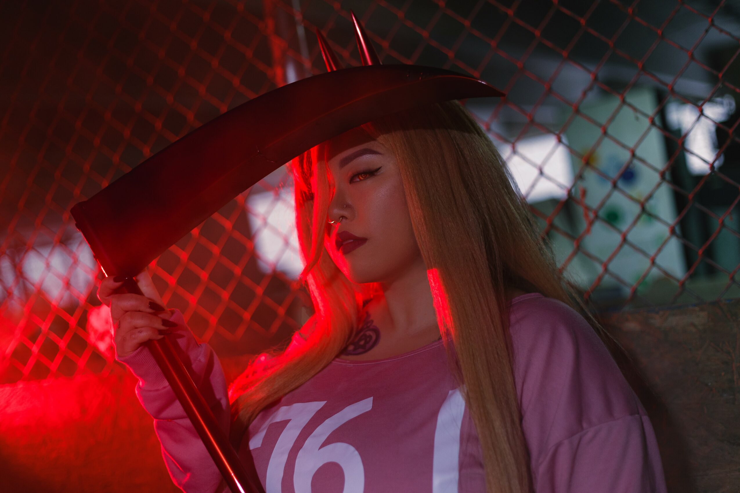 Young woman with long hair holding an umbrella, illuminated by red light, standing near a chain-link fence at night.