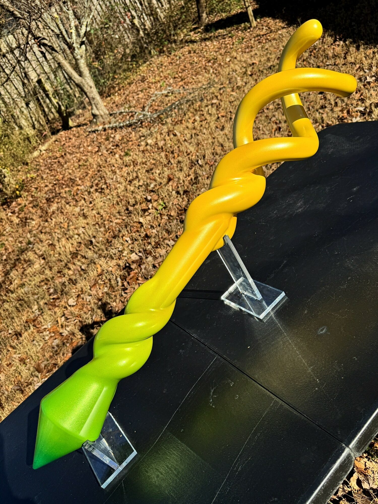 Colorful twisted sculpture with yellow and green sections on a black surface outdoors, with trees and fallen leaves in background.