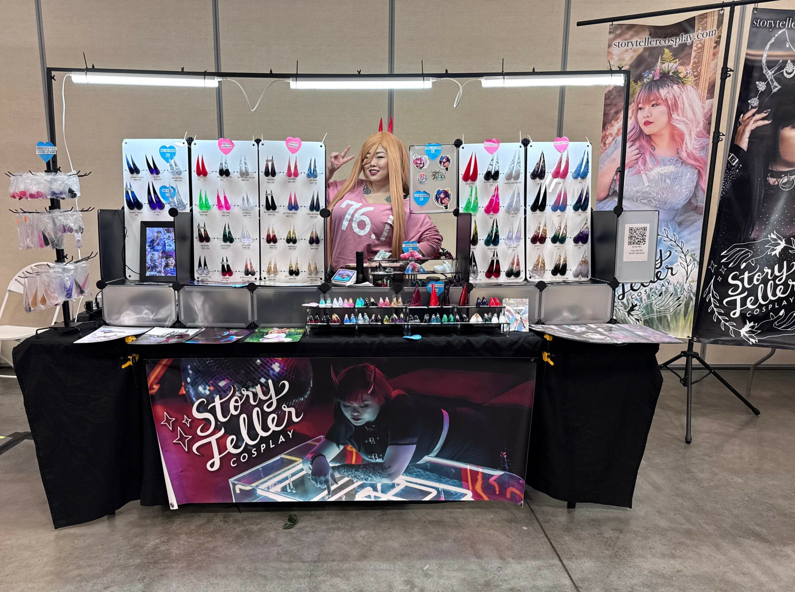 Vendor booth with colorful earrings display, a person behind the table, and promotional posters in the background.