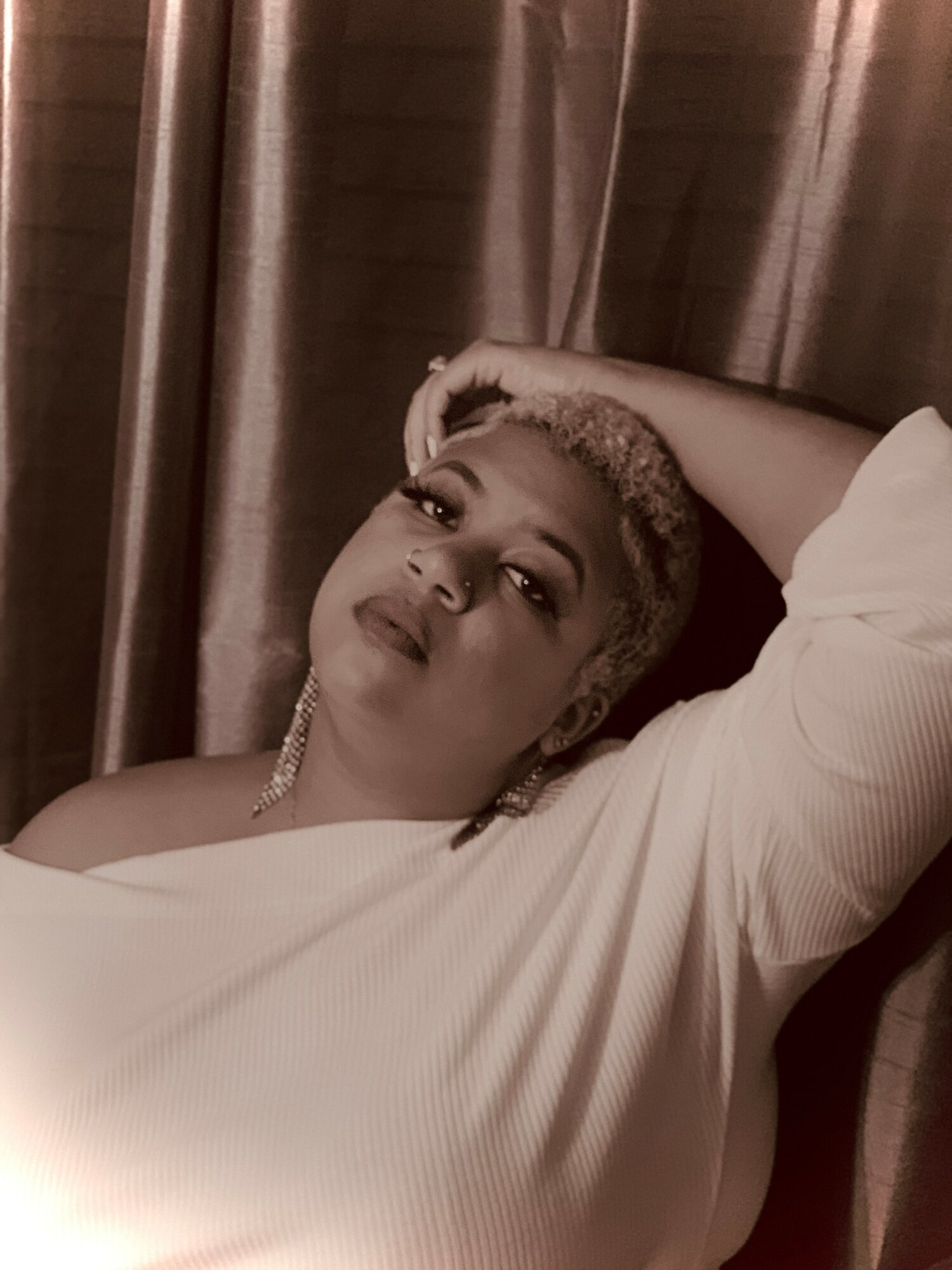 Woman with short hair and earrings, resting her head on her arm, looking at the camera, in front of curtains.