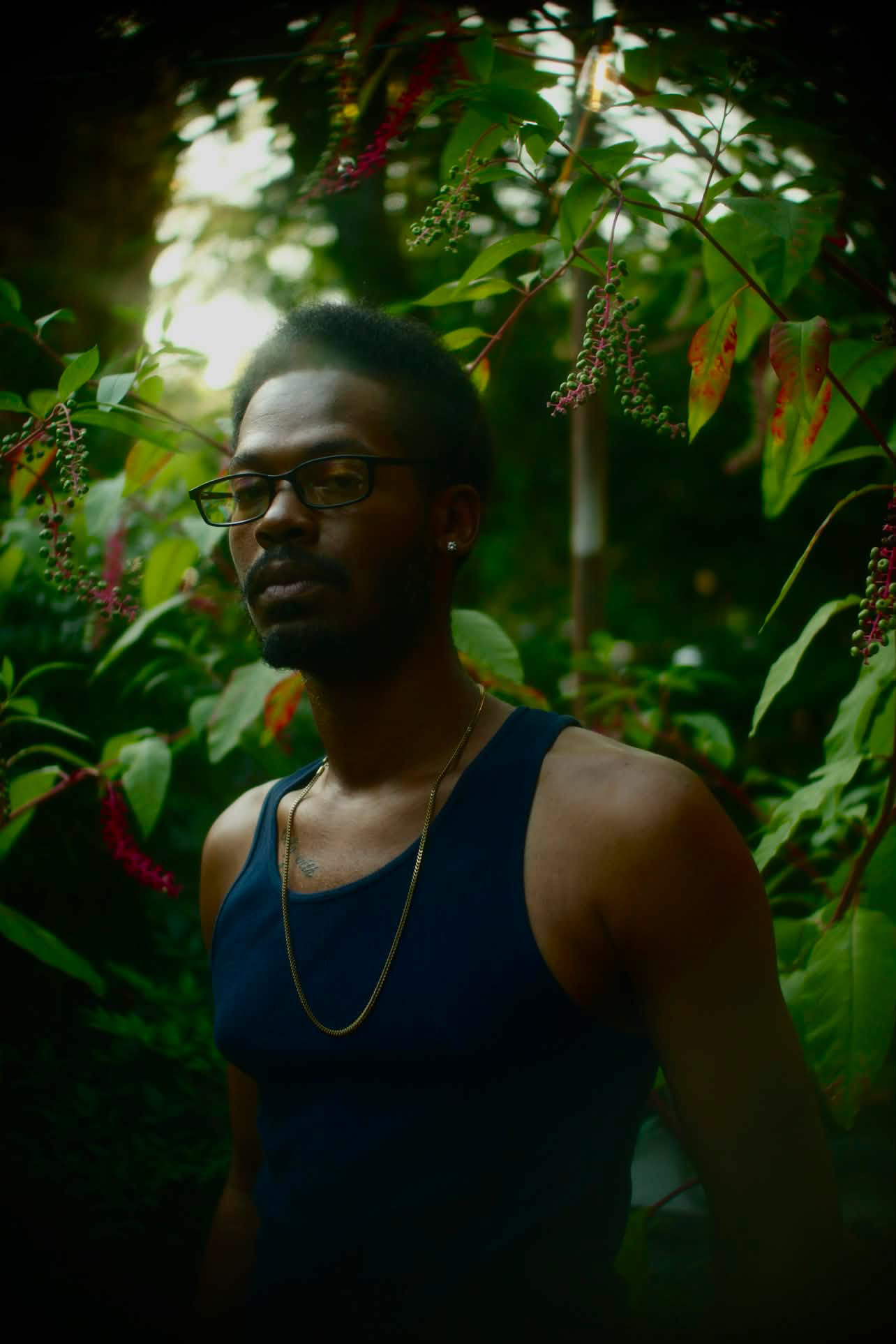 Young man with glasses and a gold chain stands among green foliage outdoors.