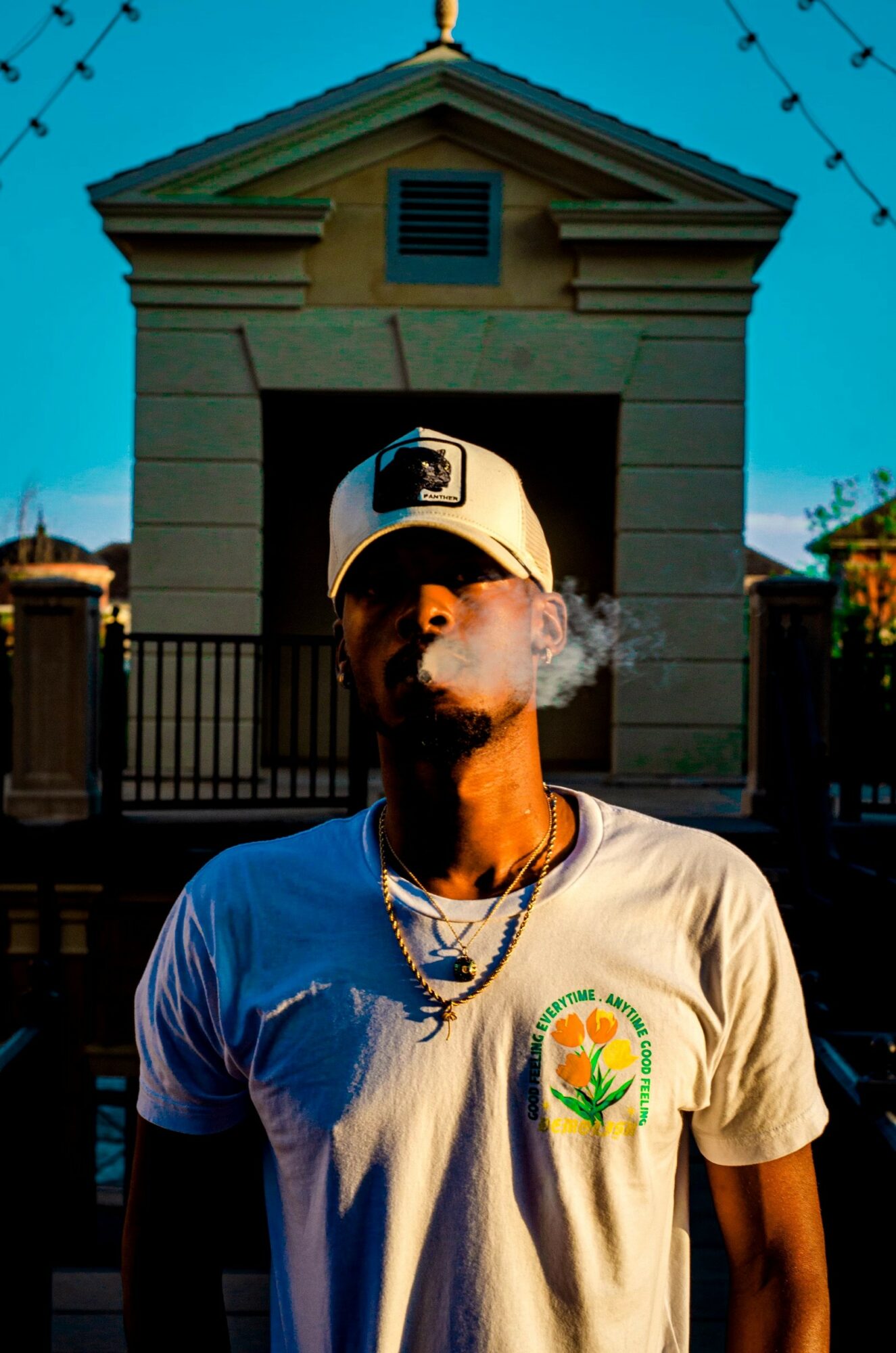 Young man wearing a cap and t-shirt with a logo, smoking, standing outdoors in front of a building with a gate.