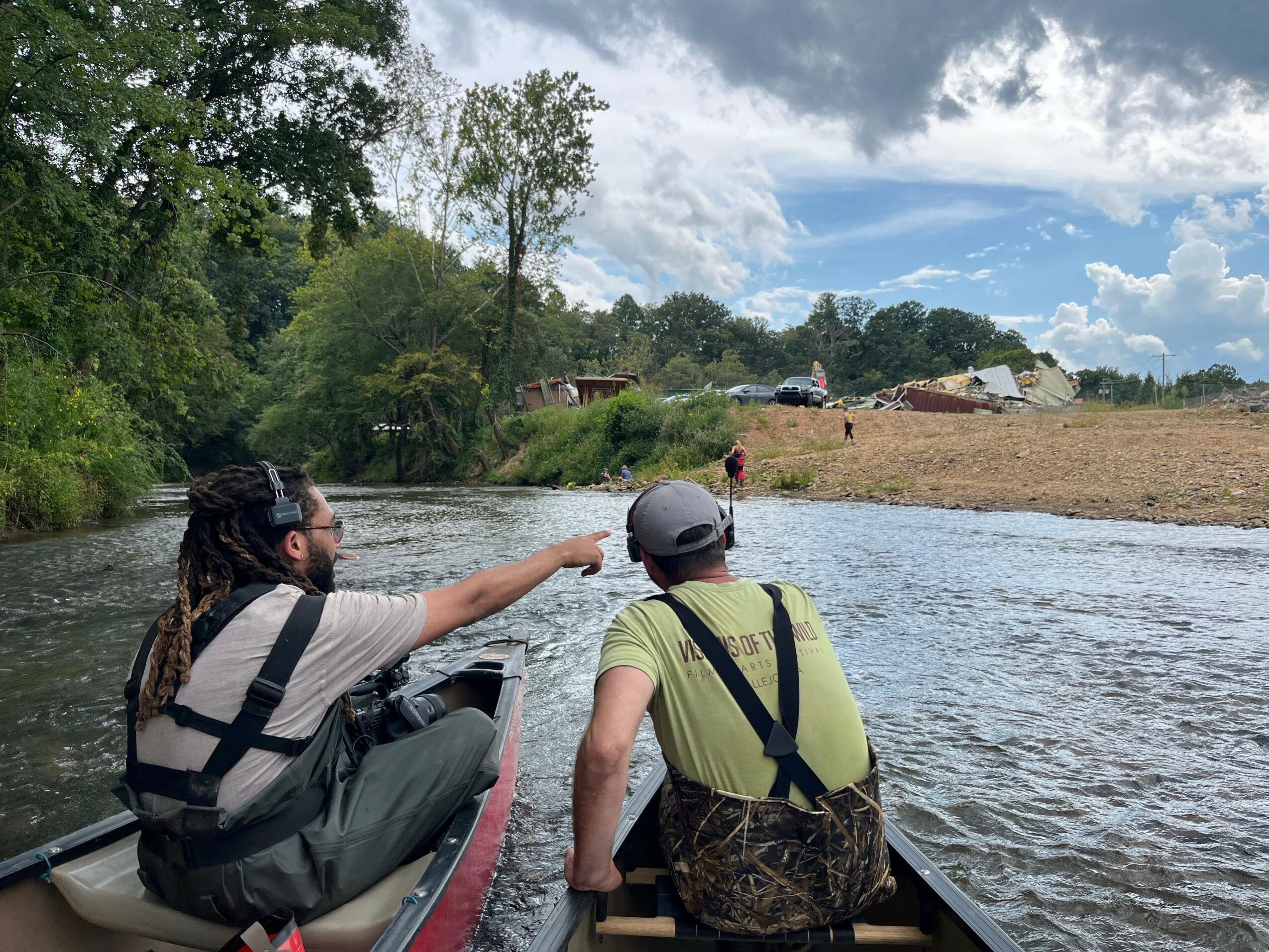 Two people sit in a boat on a river, pointing towards the opposite bank with trees and a cloudy sky in the background.