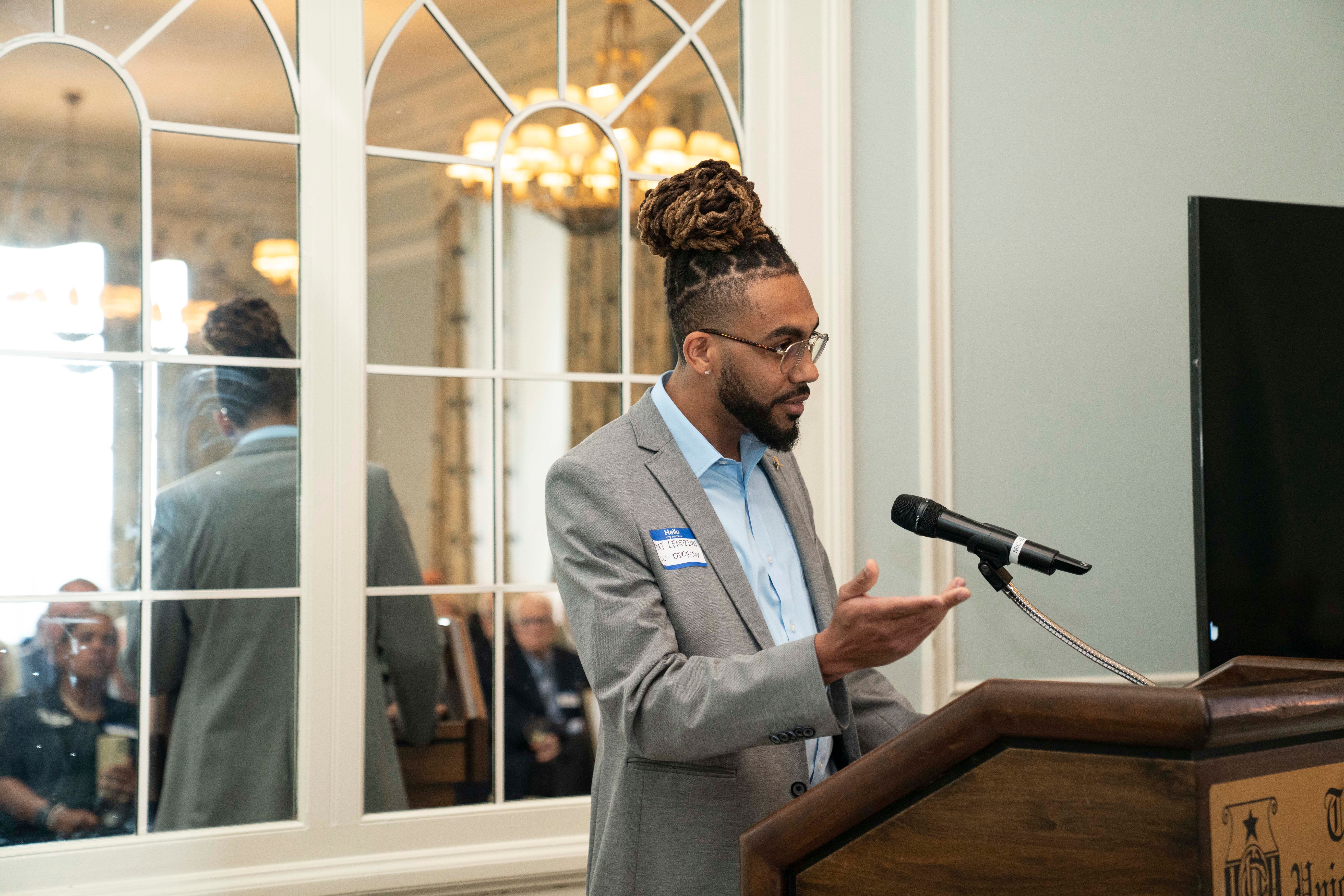 A man with glasses and a beard, wearing a gray suit, stands at a podium with a microphone, speaking or presenting.