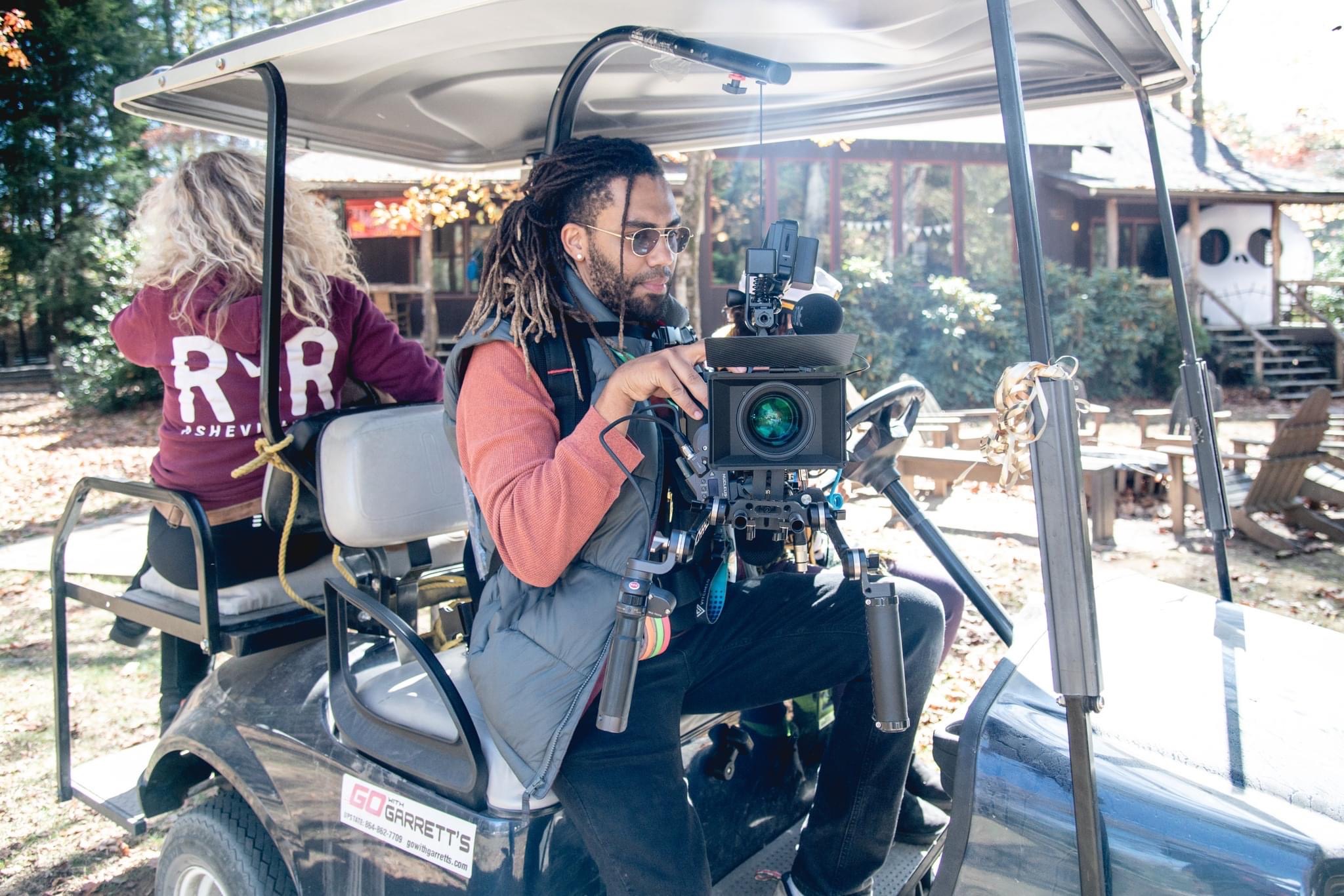 Person operating a camera on a golf cart outdoors with two women in the background, trees, and outdoor furniture visible