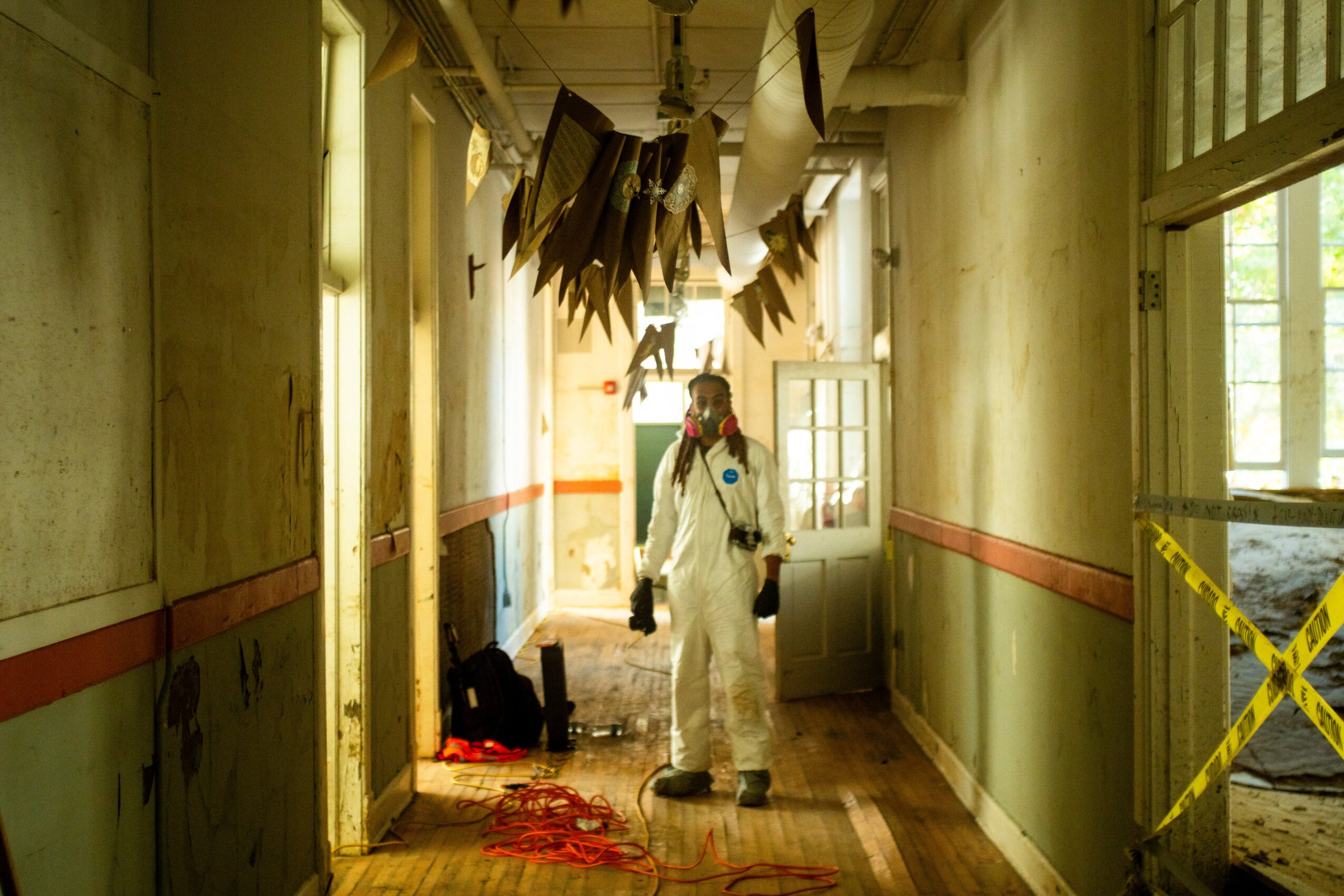 Person in protective suit and mask walking down a hallway with hanging fabric and debris, sunlight through windows.