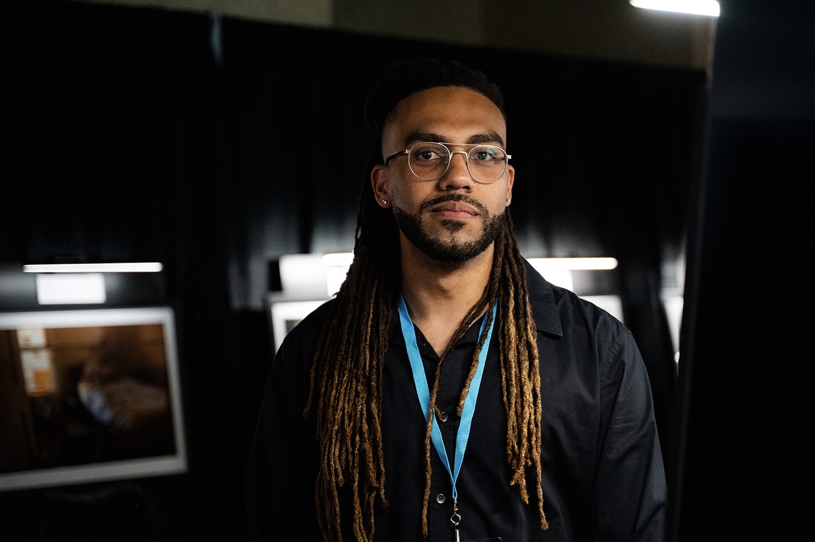 Man with glasses and long dreadlocks wearing a dark shirt and lanyard, standing indoors with screens in background.