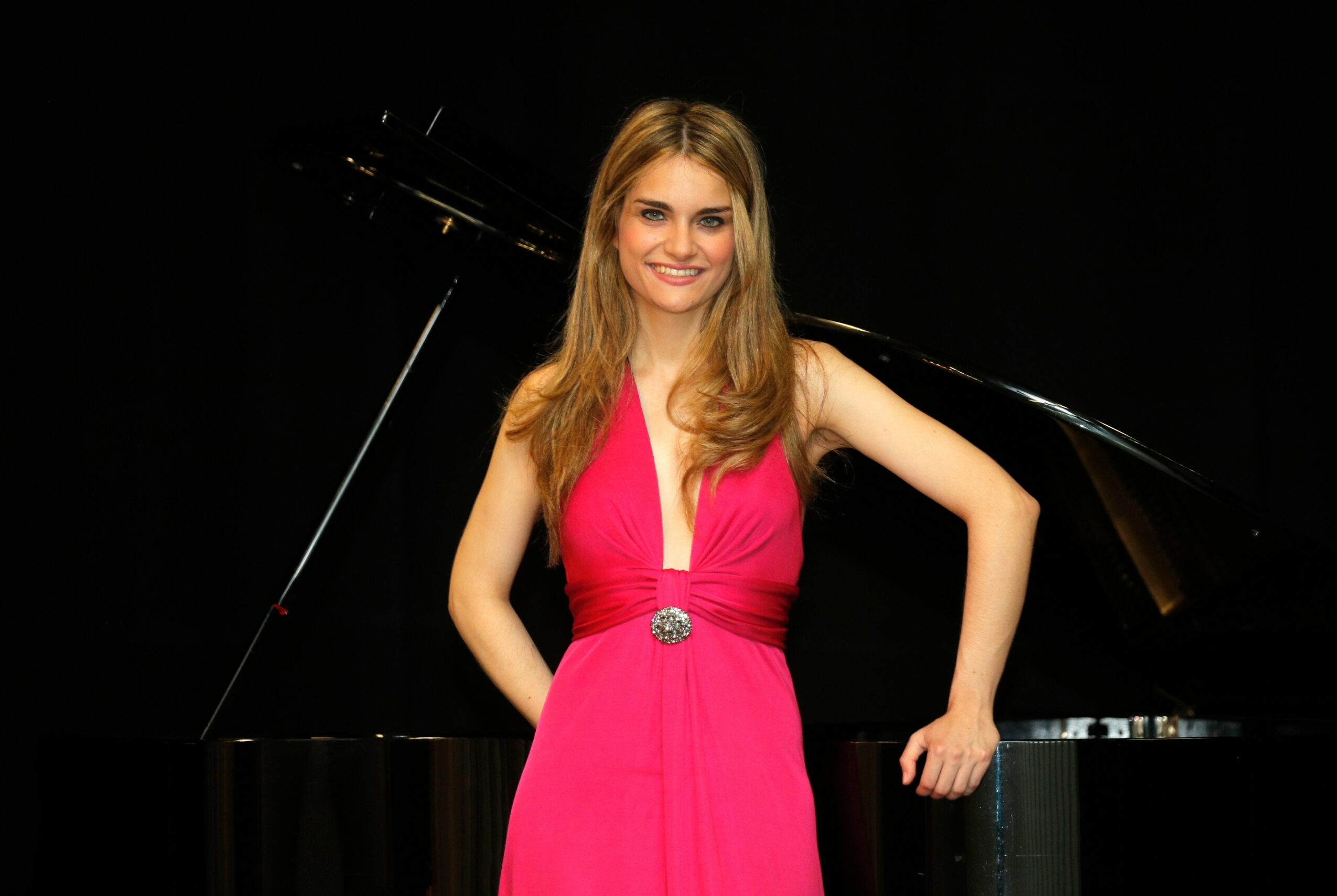 Young woman in pink dress standing in front of a black grand piano, smiling, with hands on hips.