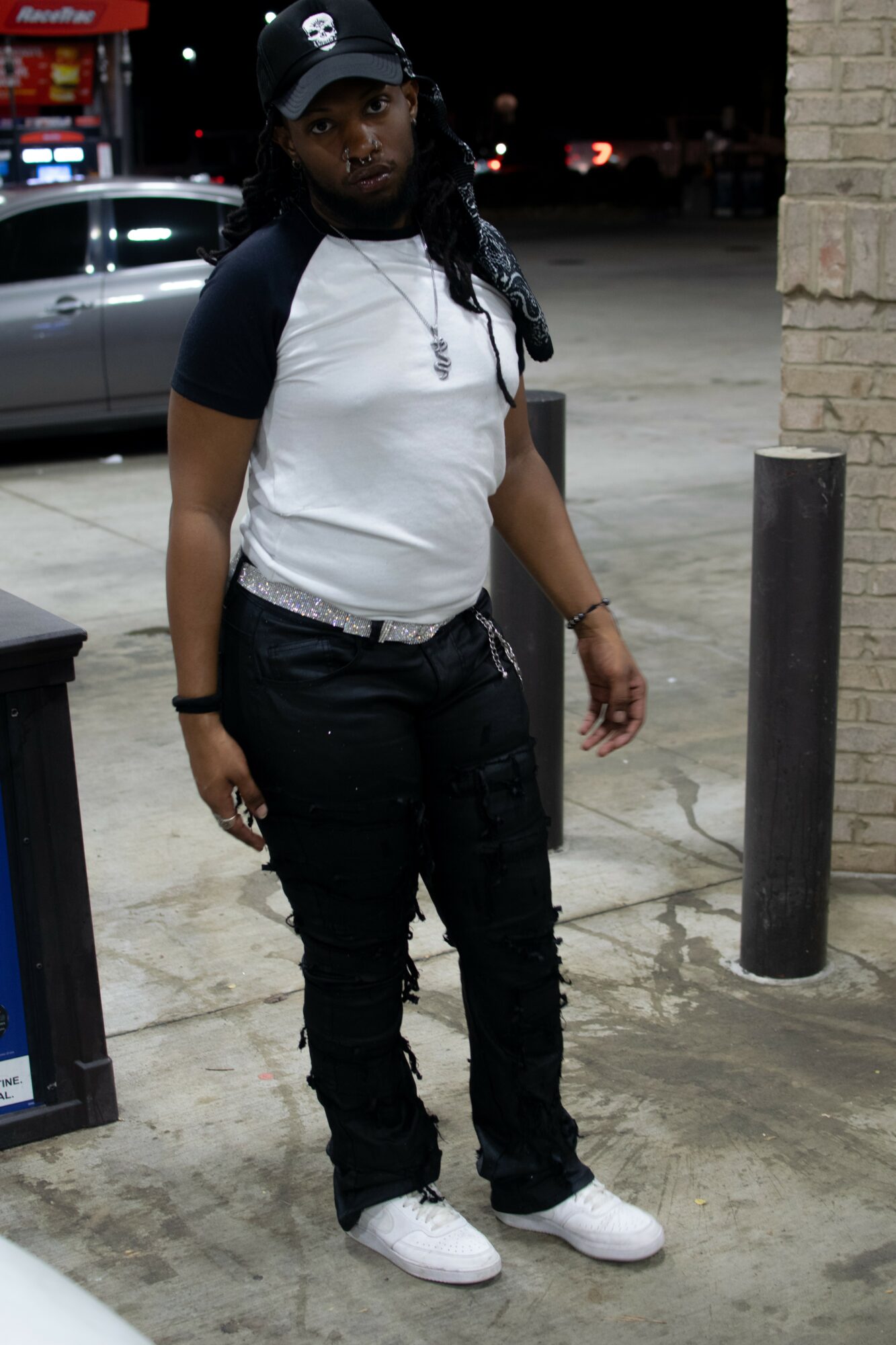 Person standing outdoors at night, wearing a baseball cap, white and black shirt, black pants, and white shoes.