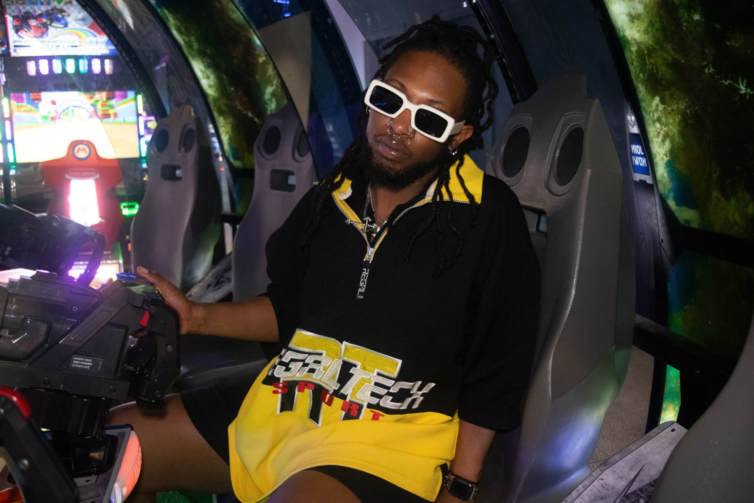 Person with sunglasses sitting inside a vehicle with a gaming console, wearing a black and yellow jacket, in a futuristic setting.