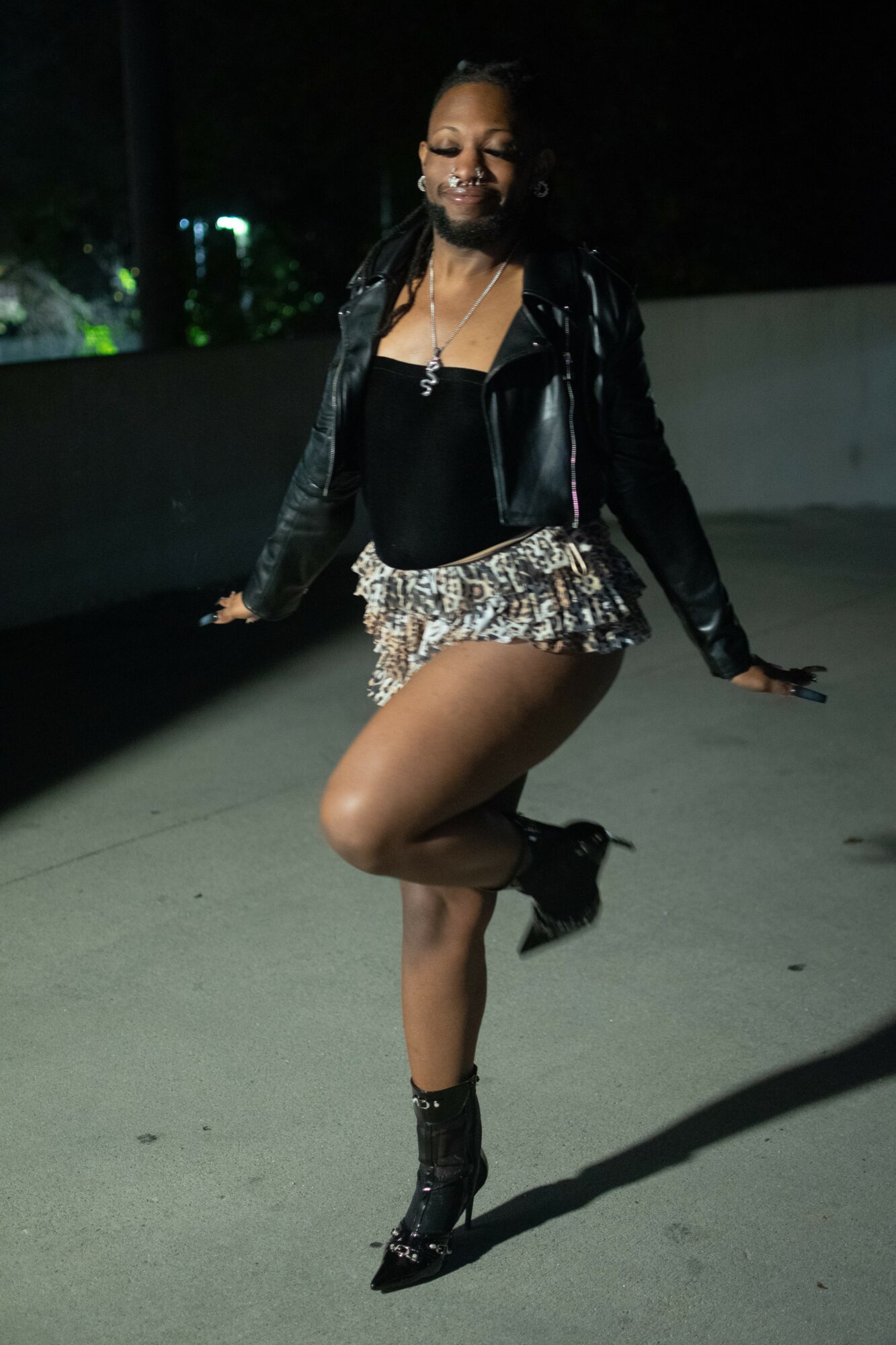 Person standing outdoors at night, smiling, wearing a black jacket, patterned shorts, and high-heeled boots, with one leg raised.