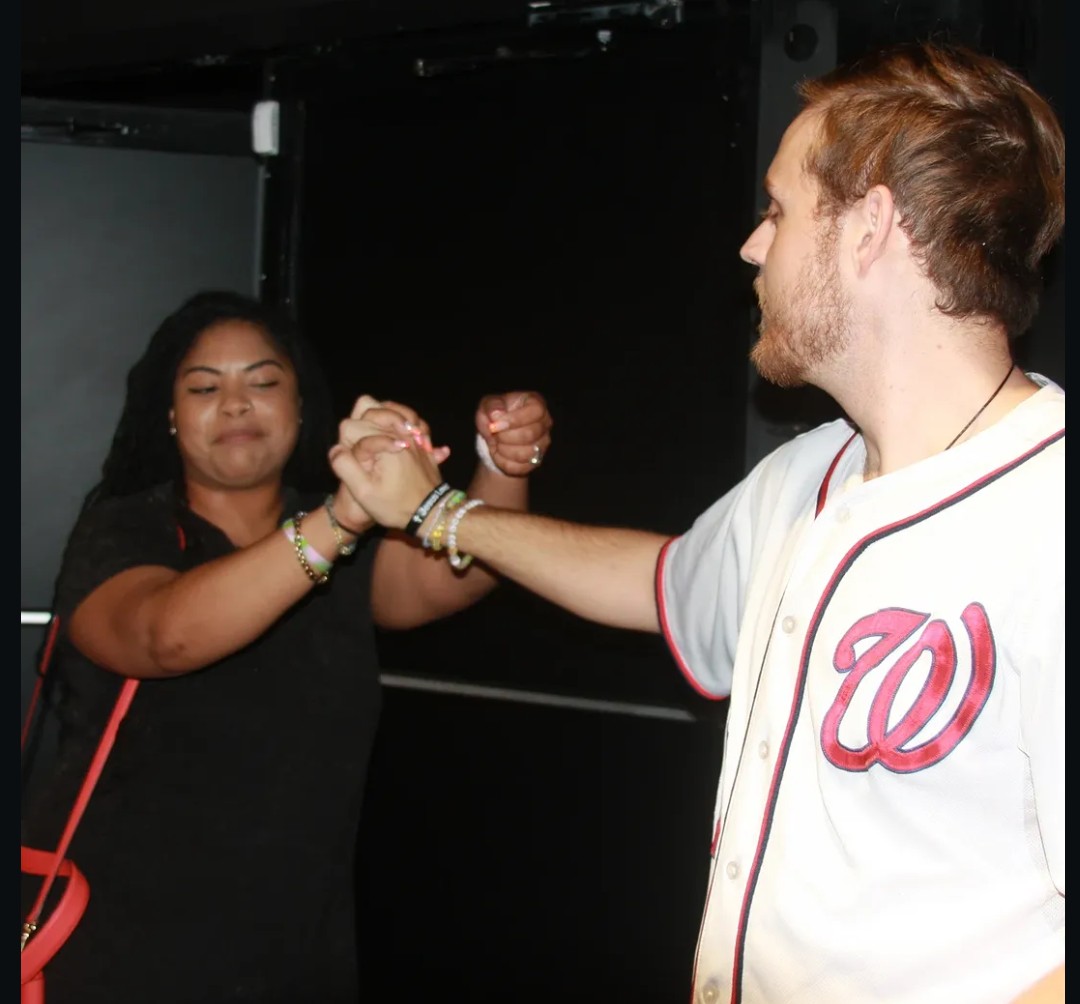 Two people, a woman and a man, shake hands and smile, in a dark indoor setting.