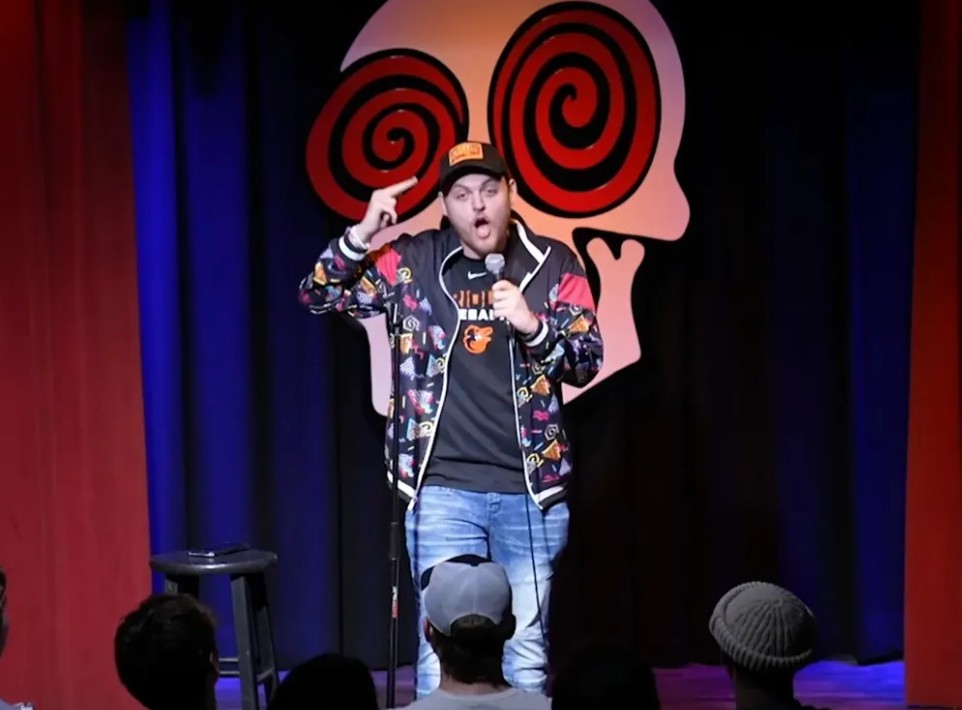 Stand-up comedian performs on stage with a colorful background, holding a microphone and gesturing with his right hand.