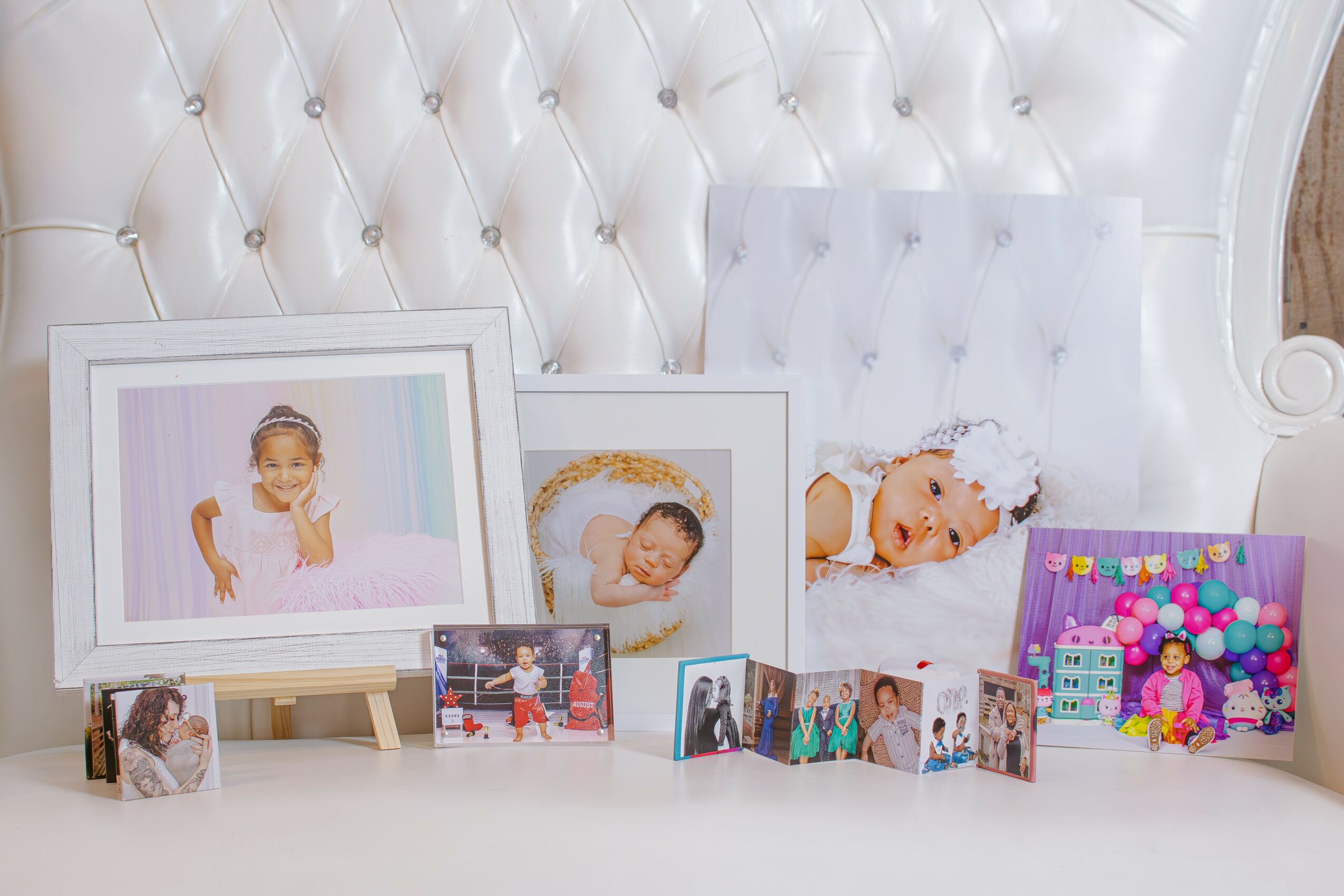 Collection of framed and unframed baby photos on a white tufted surface, with a white chair in the background.