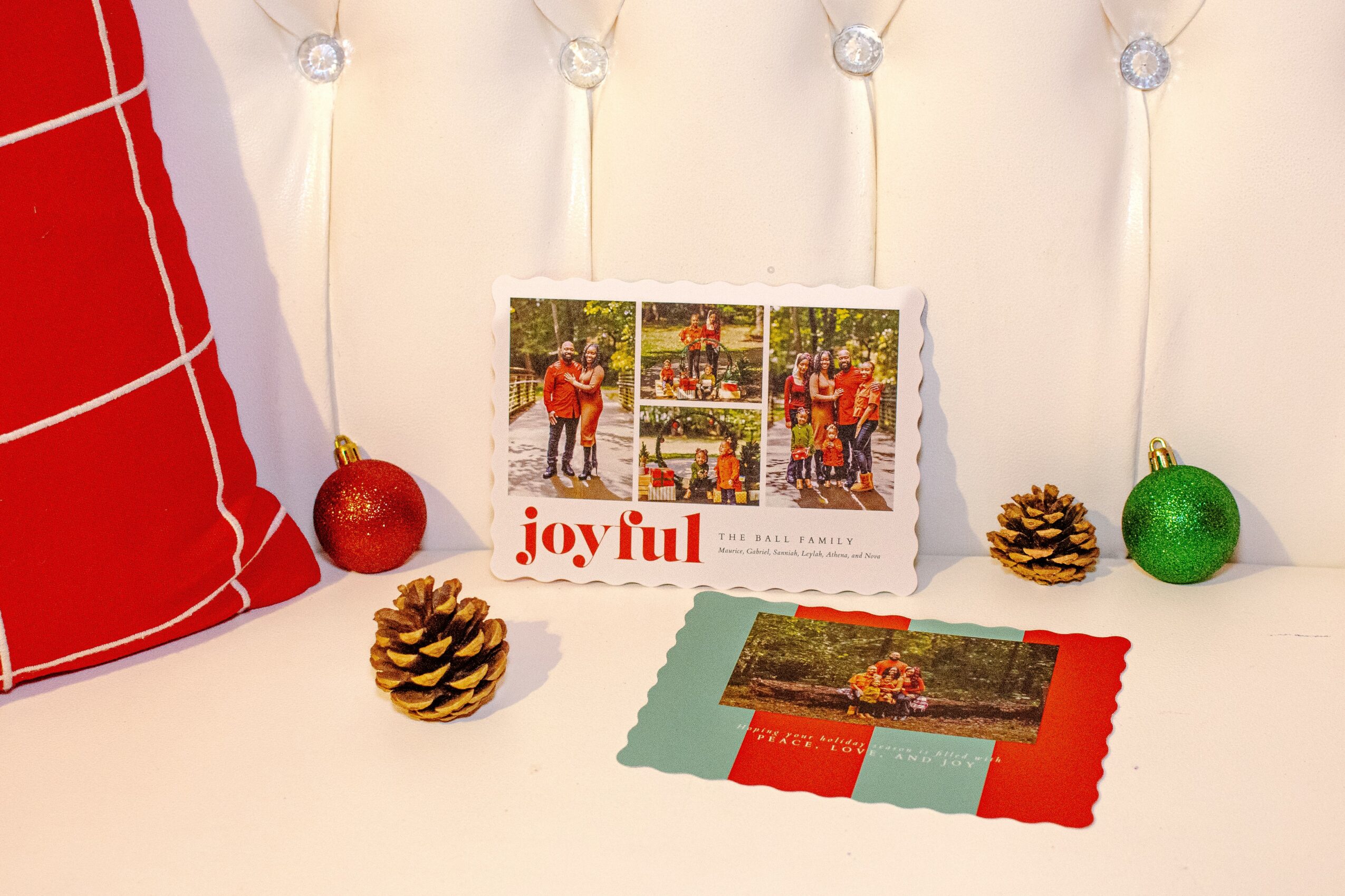 Decorative holiday scene with a photo card, pinecones, red and green ornaments, and a red pillow, on a white background.