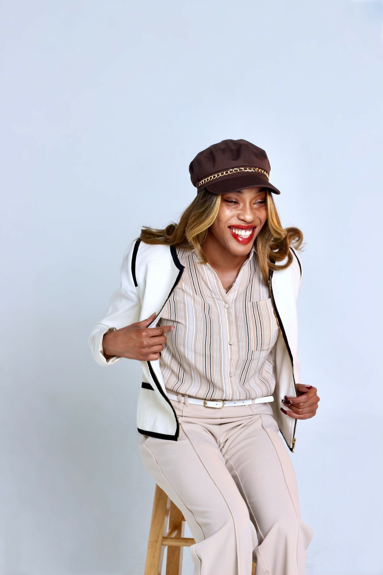 Smiling woman wearing a brown hat, white blazer, and beige outfit, sitting on a stool against a light background.
