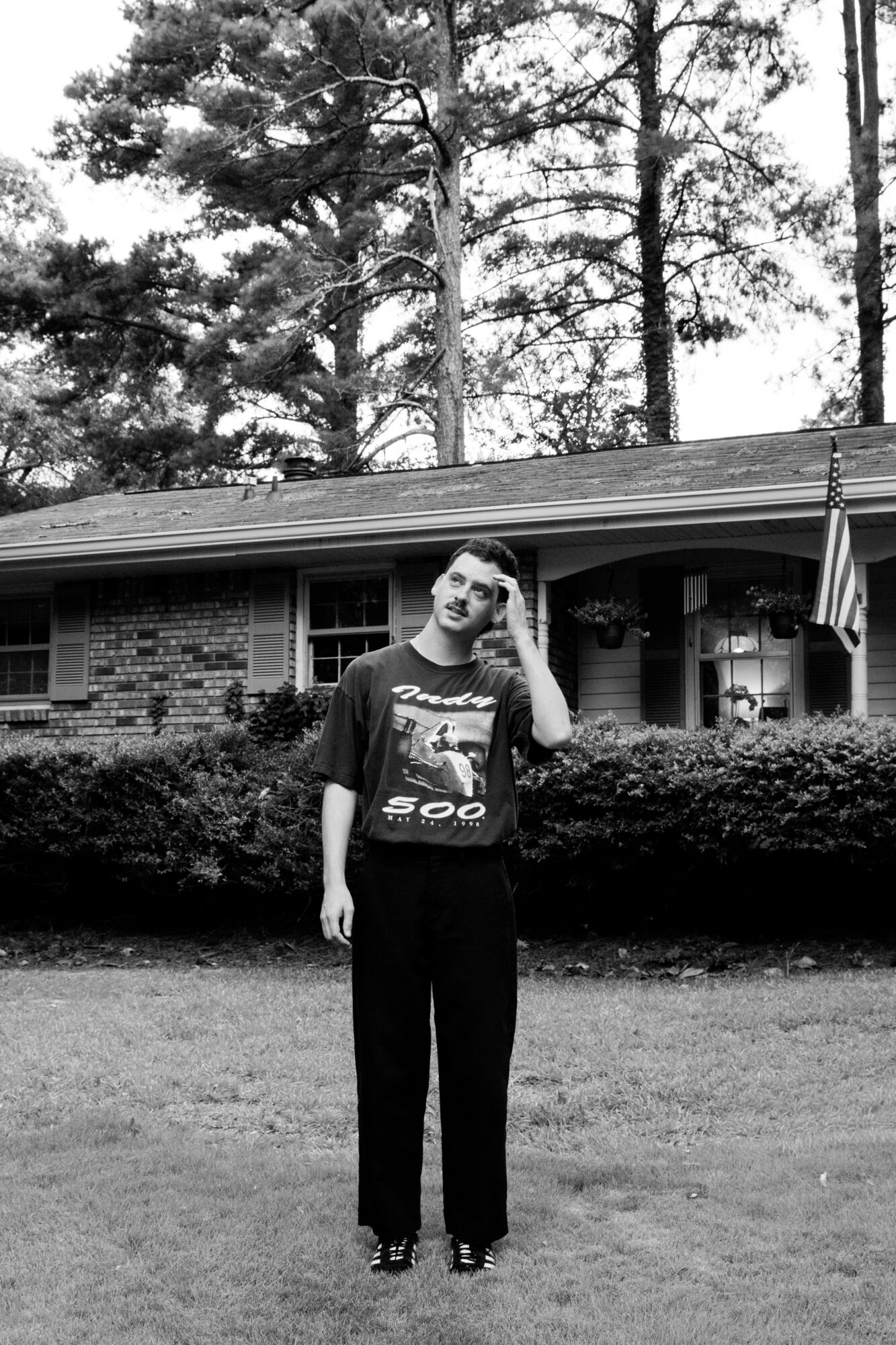 Person standing outdoors in front of a house with trees in the background, touching their head, in black and white.