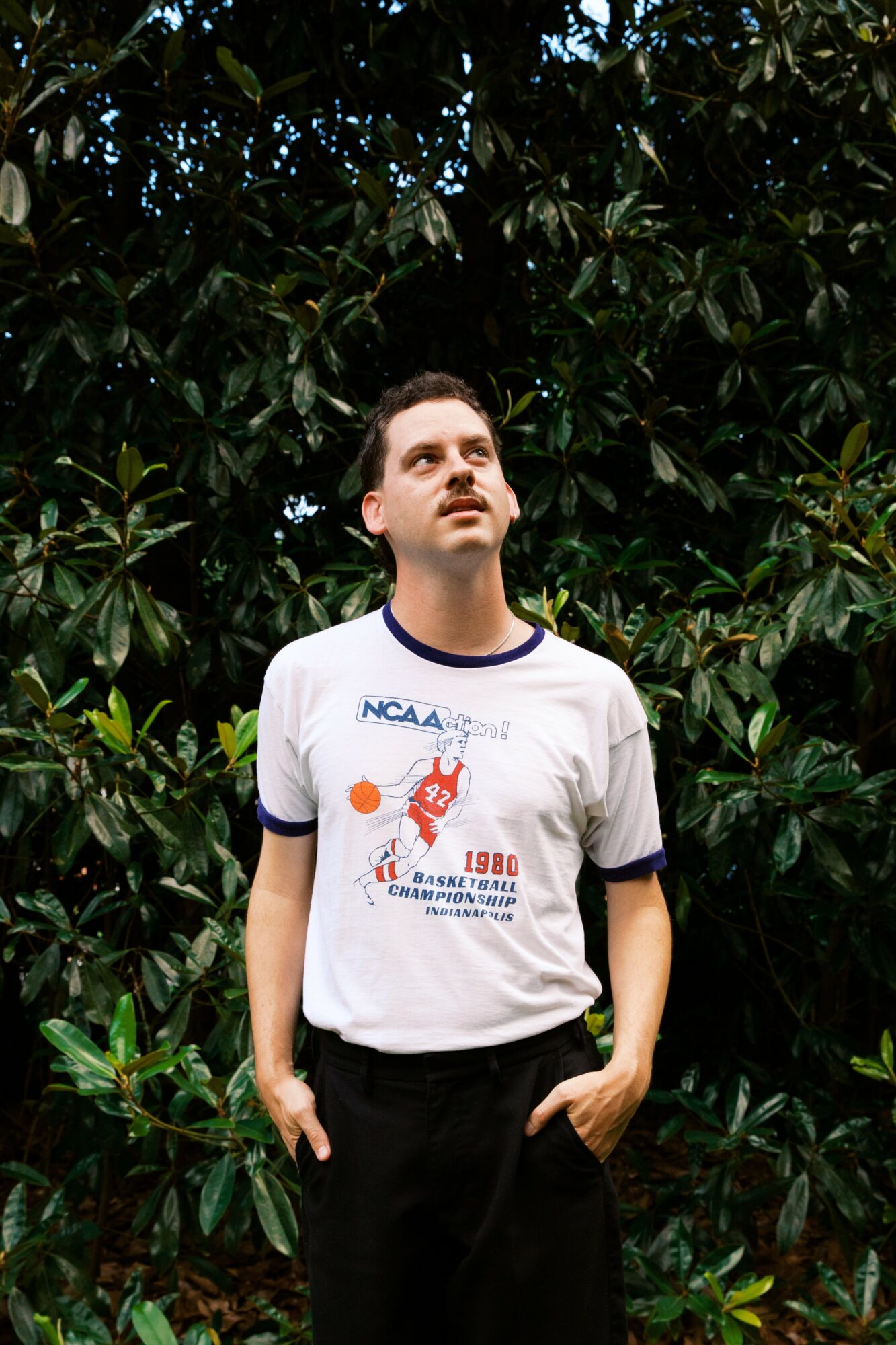 Young man with short dark hair and mustache stands outdoors in front of dense green foliage, looking upward.