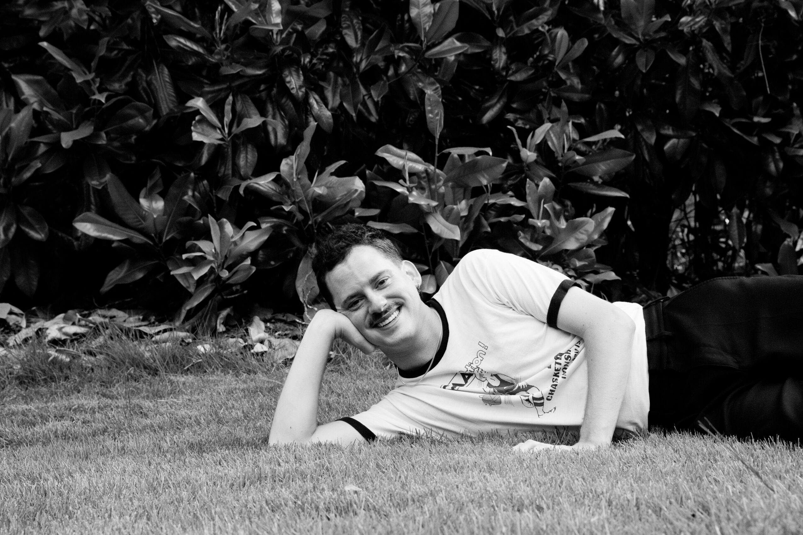 Person lying on grass, smiling, resting head on hand, surrounded by leafy plants, in black and white.