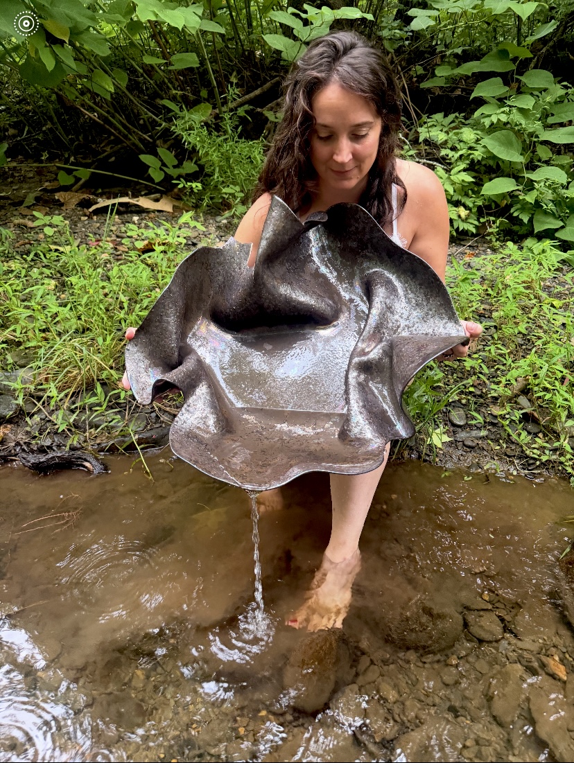 Woman holding a large, shiny, dark object in a shallow creek surrounded by green foliage.