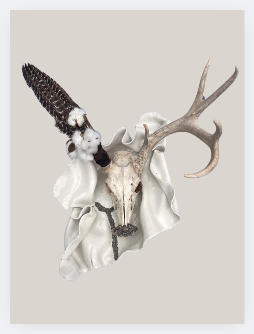 Decorative animal skull with antlers and feathers mounted on a white cloth background.