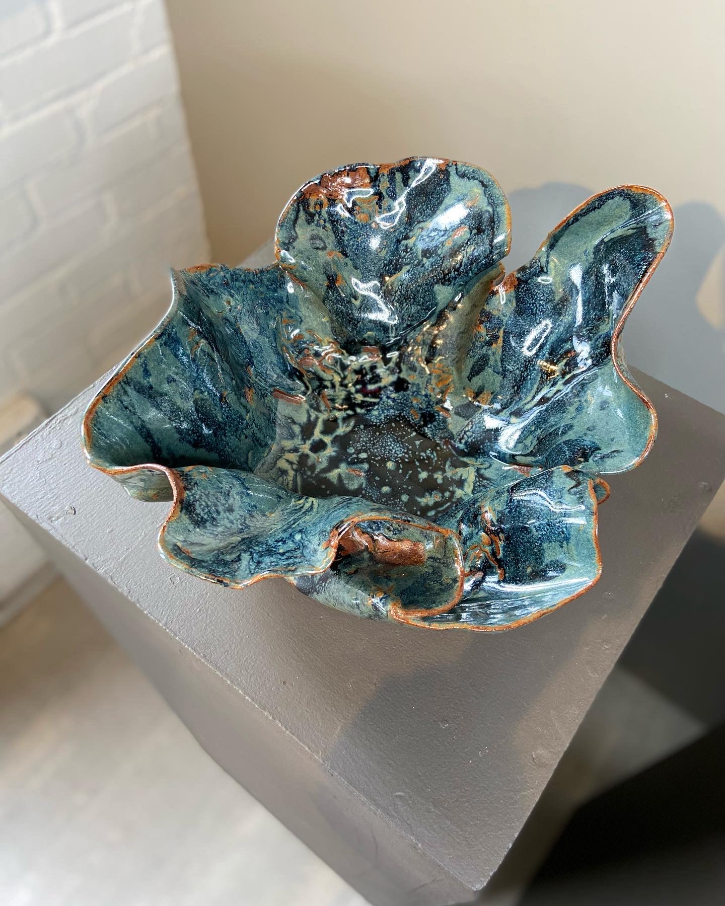 Ceramic bowl with a wavy, irregular shape, glazed in blue, green, and brown tones, on a gray pedestal.
