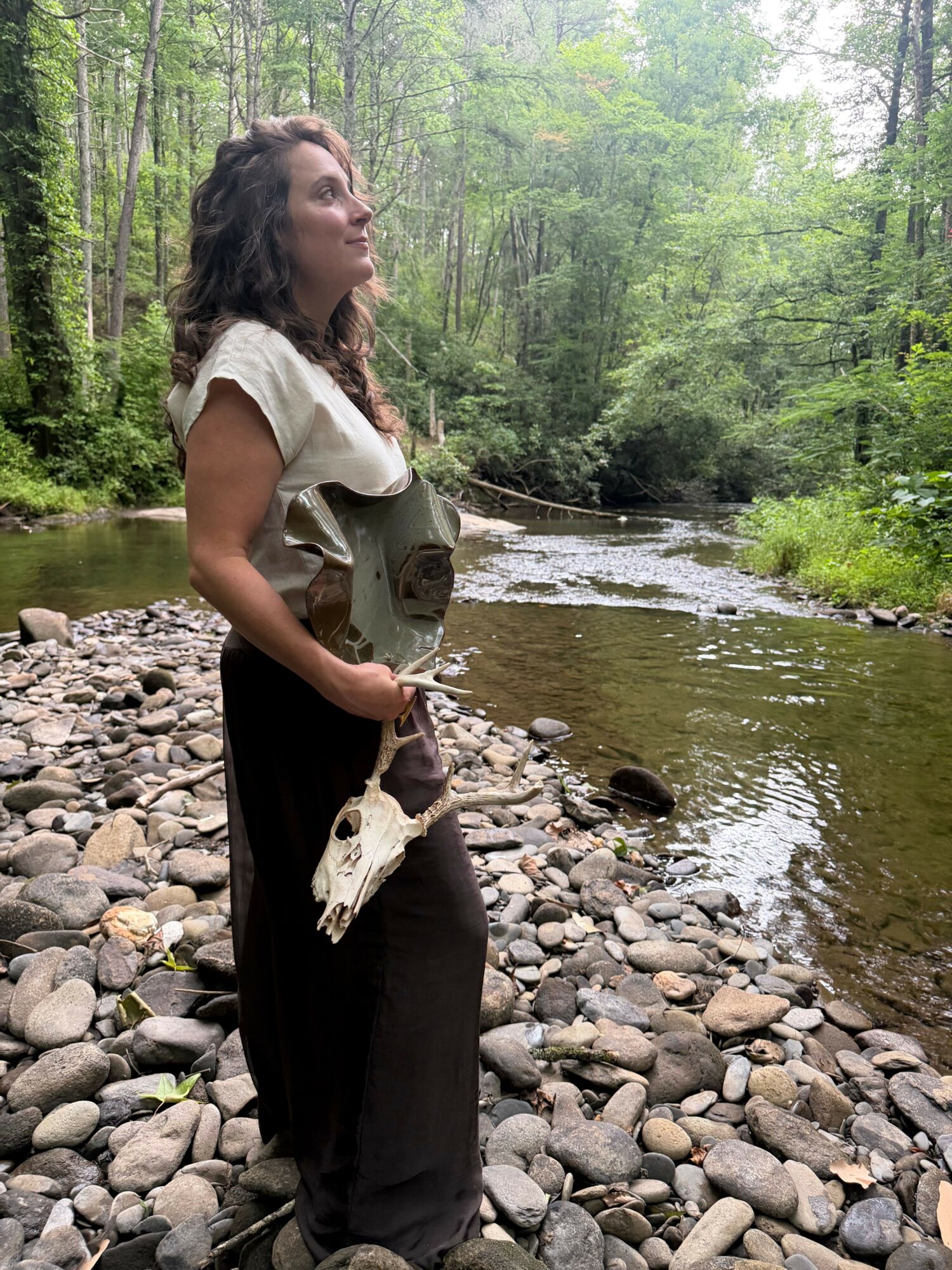 Young woman standing on rocky riverbank holding a fish, with water and trees in the background.