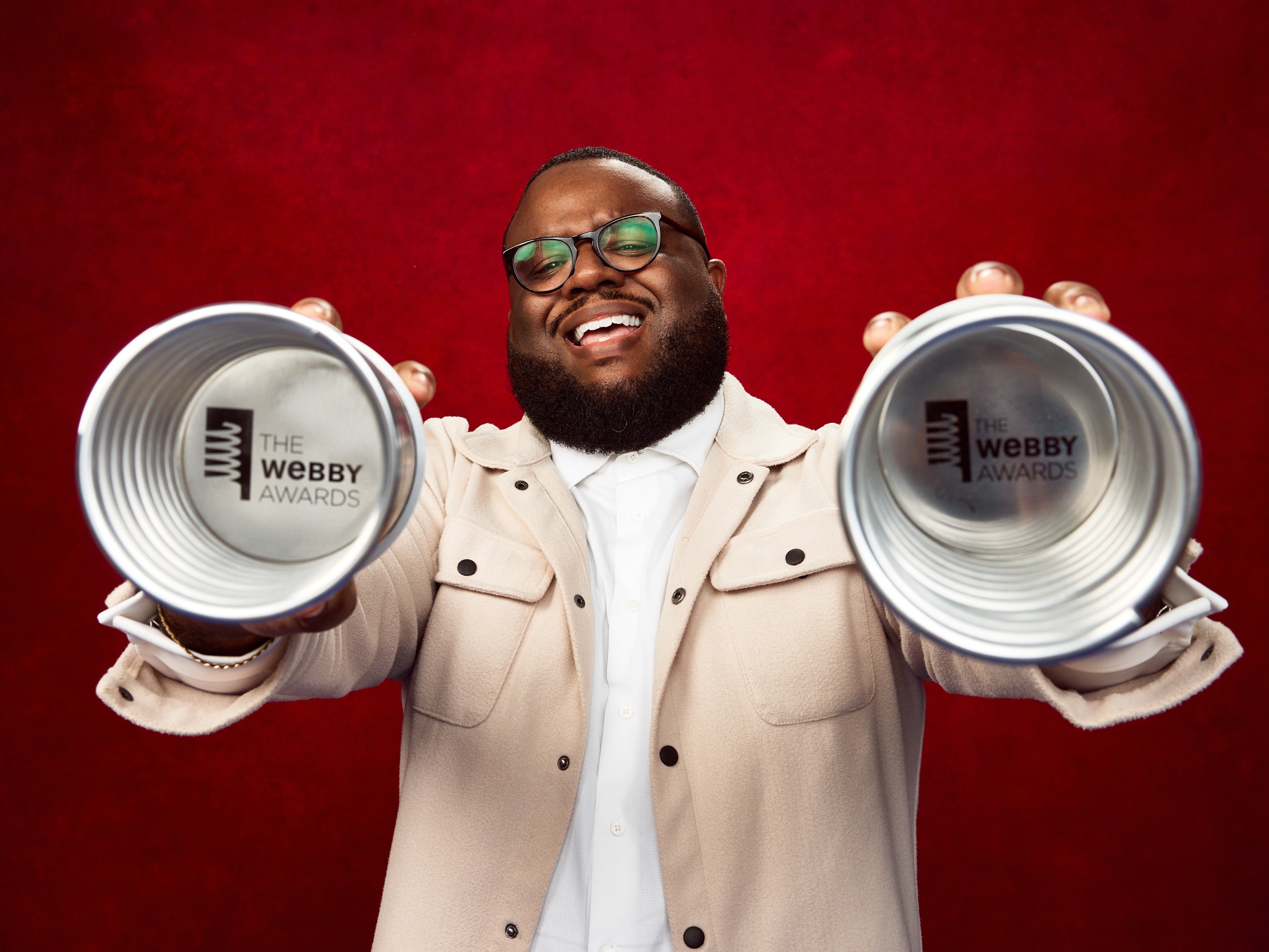 Man with glasses smiling holding two Webby Award trophies against a red background.